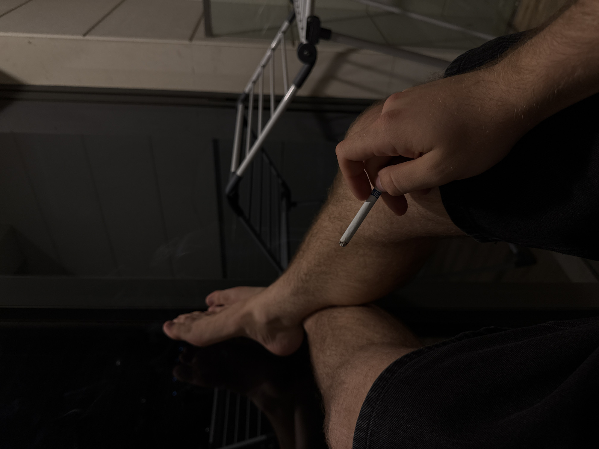 A close-up, low-angle shot shows a person's legs and hand holding a lit cigarette. The person is wearing black shorts and is seated, with their legs crossed. One foot is visible, bare and resting on the other leg. The background is dark and out of focus, with hints of a metallic structure that appears to be a drying rack. The lighting is dim, suggesting an indoor setting at night or in low light conditions. The overall mood is relaxed and contemplative. No visible text is present in the image. The location context of Dubai, United Arab Emirates, does not provide any specific visual cues within the image itself.
