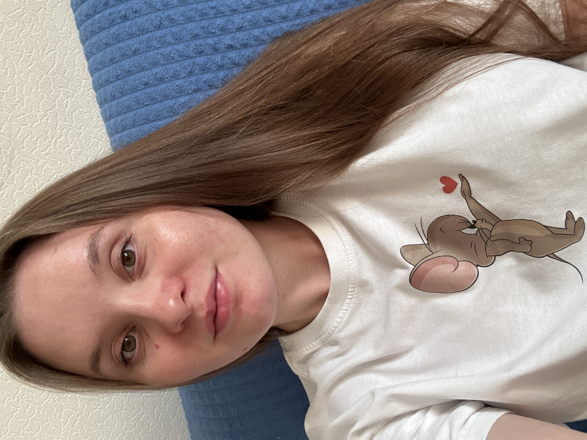 A young woman with long, brown hair and green eyes is pictured in a close-up, selfie-style shot. She is wearing a white t-shirt with a cartoon mouse character holding a red heart. The background consists of a textured wall and a blue, ribbed surface, possibly a couch or chair. The woman appears to be indoors. The image does not provide any information about the specific location within the city of Noviy, Russia.