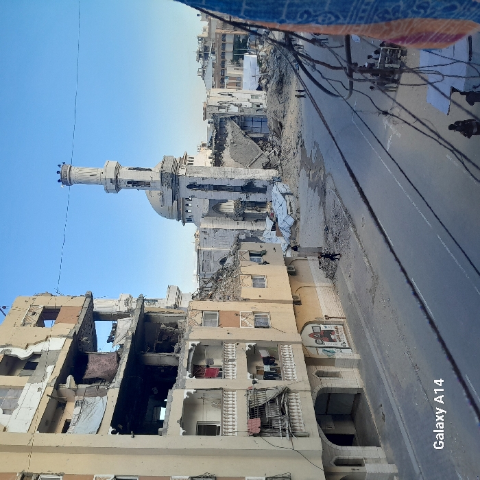 The image presents a landscape of extensive urban destruction, viewed from an elevated vantage point looking down upon a street and surrounding buildings under a clear, bright blue sky.
At the center of the scene stands a mosque, a prominent structure with a tall, slender minaret topped by a crenellated crown and a large dome. The mosque appears heavily damaged, with sections of its exterior crumbling and parts of its surface blackened, suggesting exposure to fire or explosion. Significant piles of rubble and debris are scattered around its base.
To the left of the street, in the foreground, is a multi-story residential building that has sustained severe damage. Large portions of its outer walls are missing, exposing the interiors of several apartments on multiple floors. Debris hangs precariously from damaged sections, and various fabrics or clothes are visible hanging from balconies and window openings.
A paved street runs through the middle ground, appearing relatively clear of major debris on its surface, though large mounds of rubble, dirt, and damaged building materials line its edges, especially alongside the compromised structures. Further into the background, additional buildings are visible, also showing signs of damage, indicative of a densely populated urban area.
Overhead, in the upper right portion of the image, a section of blue patterned fabric or awning is visible, along with a dense network of black electrical or communication wires strung haphazardly across the street. A few small, indistinct figures of people can be seen on the street, one near the base of the mosque and others further along, but no specific activity is clearly evident.
A notable detail on the wall of a building located on the right side of the street is a partial mural or painted image depicting what appears to be a red tractor or similar construction vehicle. The clear sky and bright illumination suggest it is daytime. The overall impression is one of widespread devastation and the aftermath of a destructive event. A watermark "Galaxy A14" is visible in the lower right corner of the image.