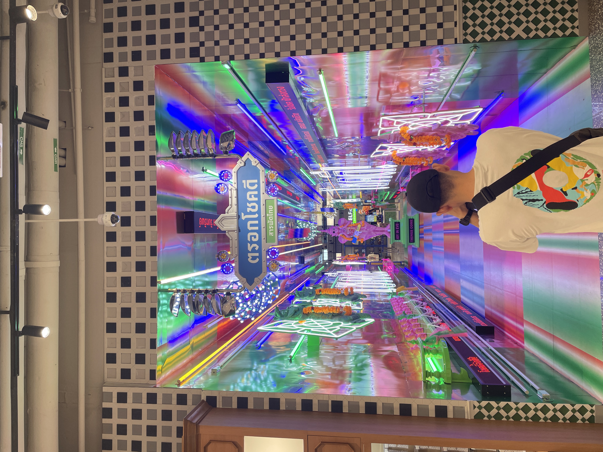 The image captures a vivid, perspective-skewed indoor scene with a person viewing an illuminated display. The individual, positioned in the right-hand side of the frame, is wearing a white shirt with colorful designs, a black hat, and a shoulder strap. Their attention is directed towards a brightly lit display that seems to stretch out like a tunnel.
The display is characterized by neon lights, signs in Thai script, and various decorative elements. The colors are intense, with prominent shades of blue, green, pink, and orange. This creates a tunnel-like effect, drawing the viewer's eye toward the end of the display.
The walls and ceiling surrounding the display are decorated with a checkered pattern in black and white, providing a stark contrast to the vibrant display. Additional elements such as surveillance cameras and lighting fixtures can be seen along the ceiling and walls.
Based on the Thai script and the cultural elements in the display, it is likely that the location is in Bangkok, Thailand. The display appears to be a decorative exhibit or installation, possibly in a shopping center or public space.