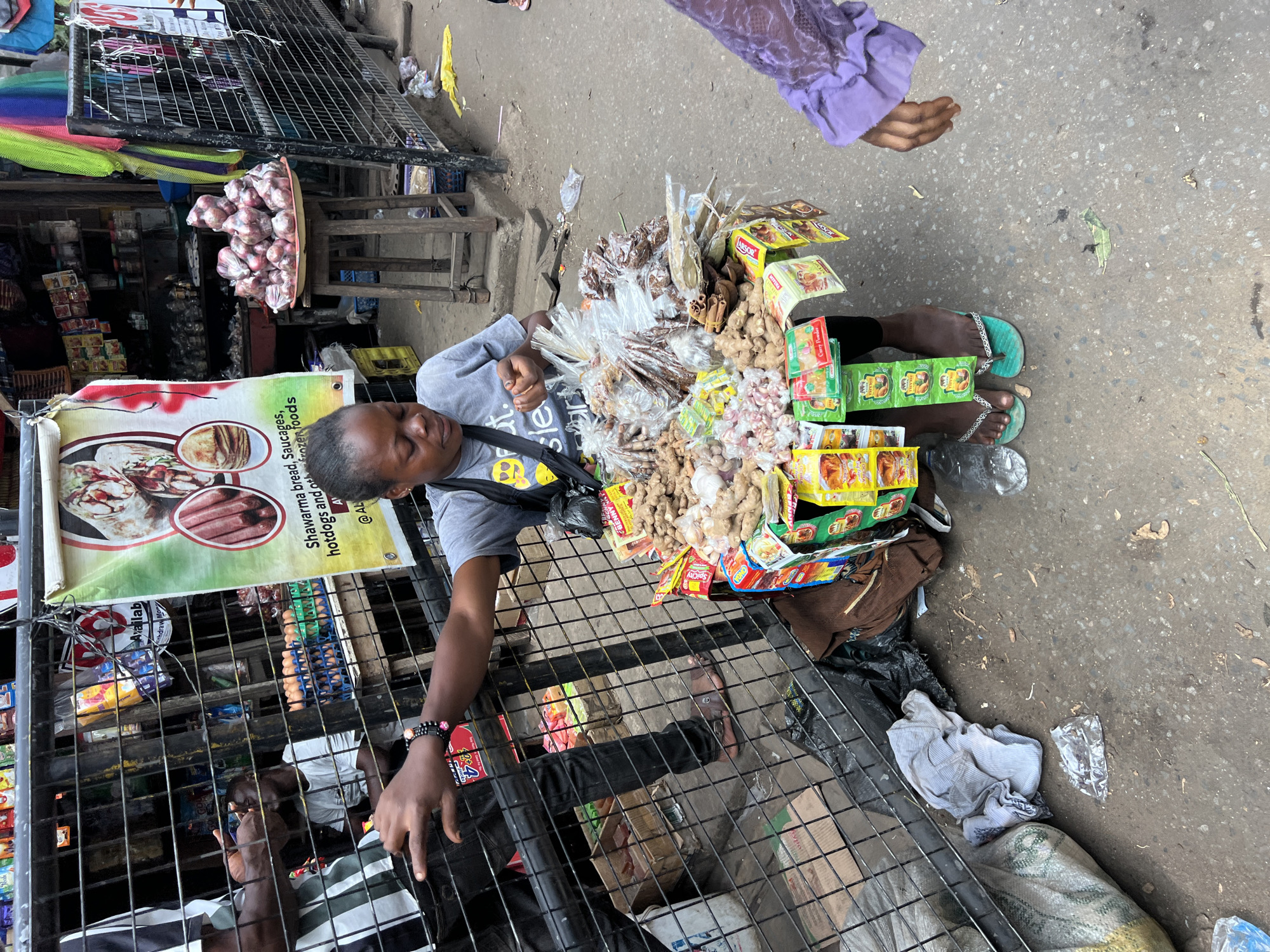 **Location:** The image is set in Nkpor, Nigeria.
**Subjects:** A street vendor is the main subject.
**People:** The vendor is a dark-skinned person wearing a gray t-shirt. Part of another person's arm, clad in a purple, long-sleeved garment, appears in the upper right corner. A second person is partially visible inside the store on the left of the street vendor.
**Objects:** The vendor is surrounded by merchandise, including packages of spices like curry powder and what appears to be ginger and other ingredients wrapped in clear plastic bags. There is a metal grill that separates the vendor from the street. Other visible items include foodstuff and eggs for sale at the location, as well as advertising posters/signs. There are also bundles of garlic.
**Environment:** The image shows an outdoor street setting in Nkpor, Nigeria. The ground is paved. There is street debris.