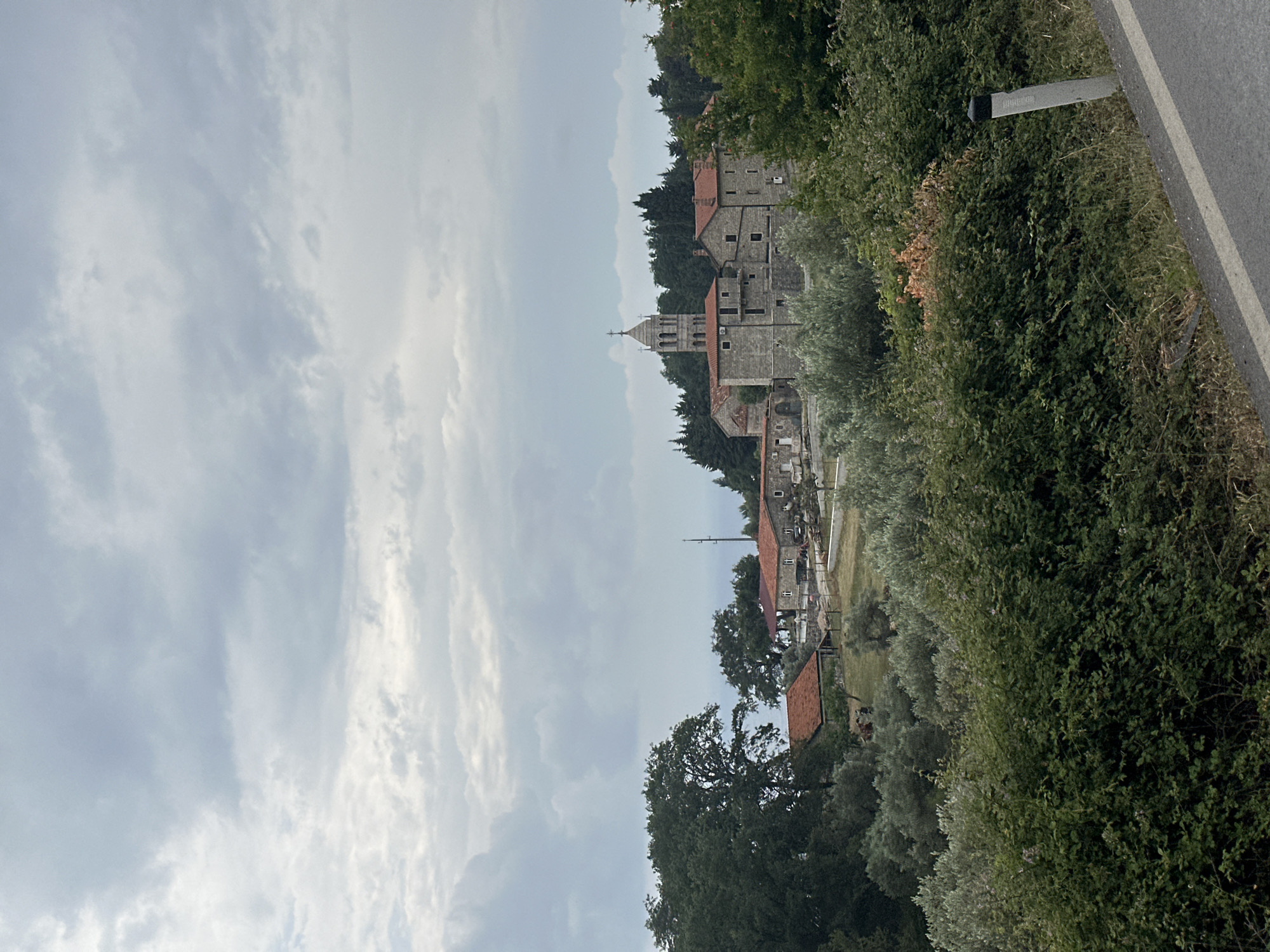 The image captures a serene, overcast daytime view of a village, likely Gornji Katun in Montenegro, nestled within a verdant, hilly landscape. The scene is dominated by traditional stone buildings, many featuring distinctive red-tiled roofs, set amongst dense green foliage.
A prominent stone church, characterized by its bell tower topped with a cross, stands out among the structures. The buildings appear to be old and well-integrated into the natural surroundings. No people or overt activities are visible, contributing to the tranquil atmosphere.
The foreground and sides of the image are filled with lush trees and thick bushes, including what appear to be olive trees. On the right, a portion of an asphalt road is visible, featuring a white lane marking. Alongside the road, a grey roadside marker stands, bearing some indistinct text that is not clearly legible.
The sky is largely covered by scattered, light grey clouds, indicating an overcast day with soft, even lighting, suggesting either late morning or early afternoon. The overall impression is one of peaceful rural life, showcasing the region's traditional architecture and natural beauty.
