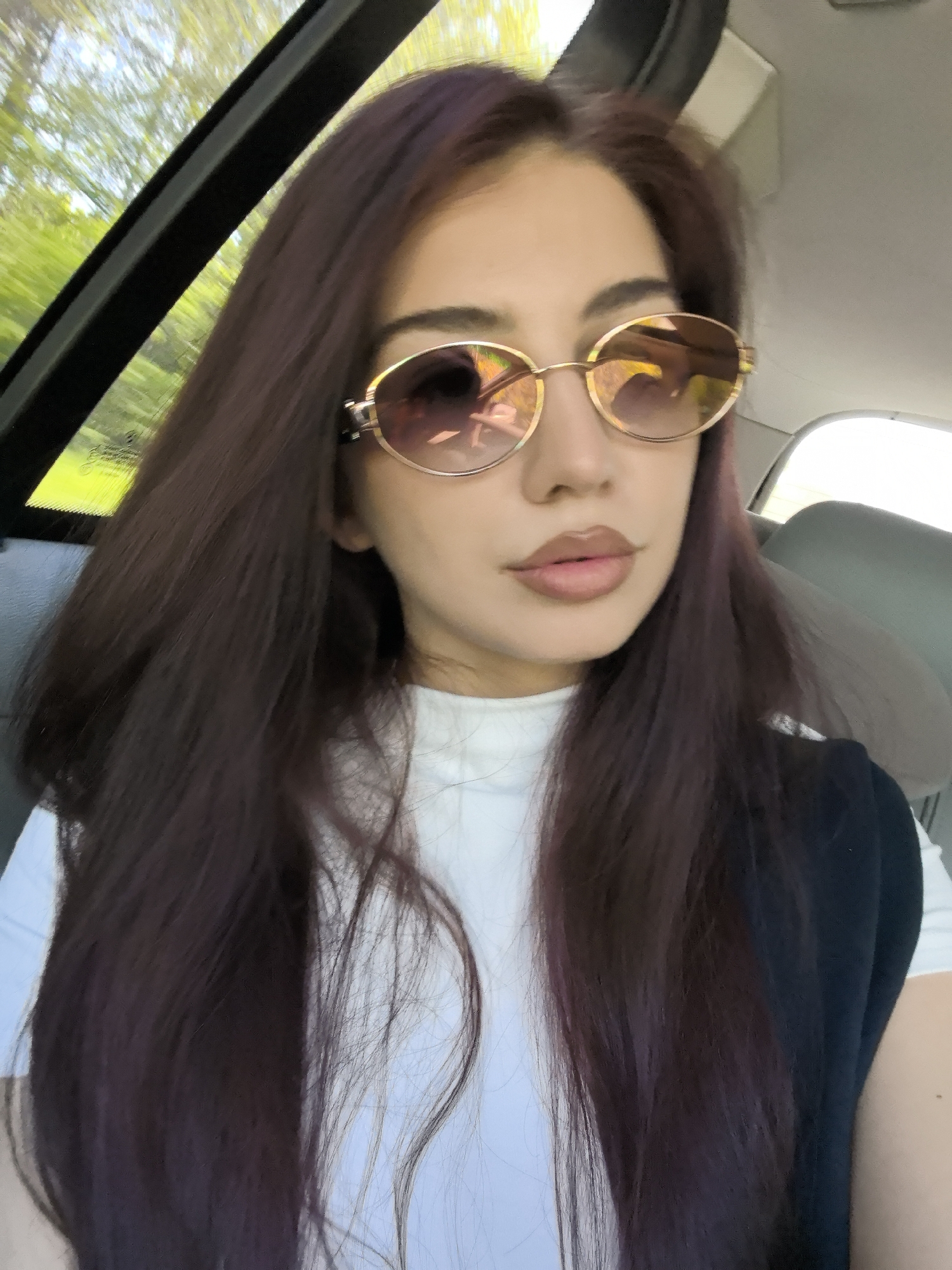 A woman with long, dark auburn hair is wearing sunglasses and a white turtleneck shirt with a dark vest. She is inside a car. The location, Bogomilovo, Bulgaria, cannot be verified from this image.