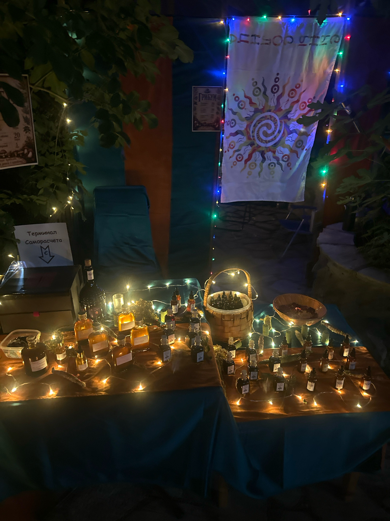 A dimly lit outdoor stall in Cetinje, Montenegro displays an assortment of small glass bottles filled with amber-colored liquid. The bottles are arranged on two tables covered with brown and teal fabric, illuminated by strings of fairy lights. Behind the tables, a white banner with a sun-like graphic and Cyrillic lettering hangs, framed by colorful LED lights. To the left, a sign with Cyrillic text reading "Терминал Саморасчета" (Self-Checkout Terminal) is visible. A wooden box and a glass bottle are also present on the left table. A woven basket filled with more small bottles sits on the right table, next to a shallow wooden bowl containing dried organic matter. Green foliage is visible in the background, suggesting an outdoor setting, possibly a market or festival. There are no people visible in the image.