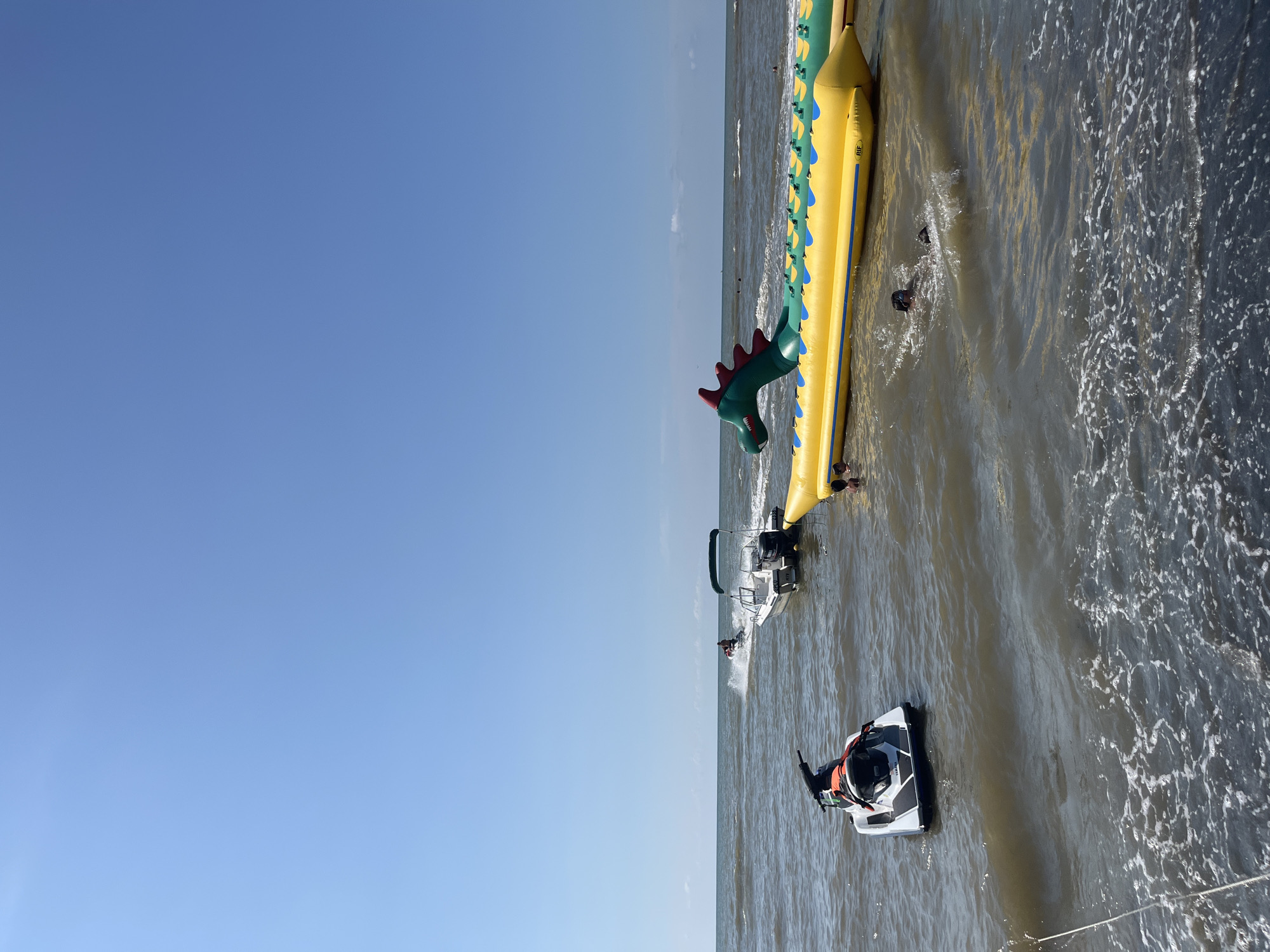 In Ureki, Georgia, on a sunny day with a clear blue sky, the choppy, brown water of the sea is populated with various recreational watercraft and inflatable attractions. In the foreground, a white and black jet ski is stationary near the shore. Further out, a white motorboat with a green canopy is moving and creating a wake. Another jet ski is further in the distance, also creating a spray. A large, yellow inflatable tube shaped like a segmented worm with a green dragon head is floating in the water, with several people swimming or wading around it. The scene depicts a typical summer day at a seaside resort.