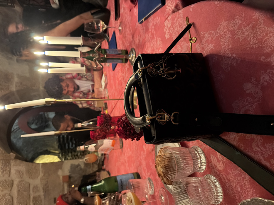 A restaurant table setting with a black Lady Dior handbag as the central focus. The table is adorned with a red patterned tablecloth and features several wine glasses, water bottles, and candles. People are seated around the table, suggesting a social gathering. The lighting is dim, creating a warm, intimate ambiance. Some architectural details of the restaurant are visible in the background.