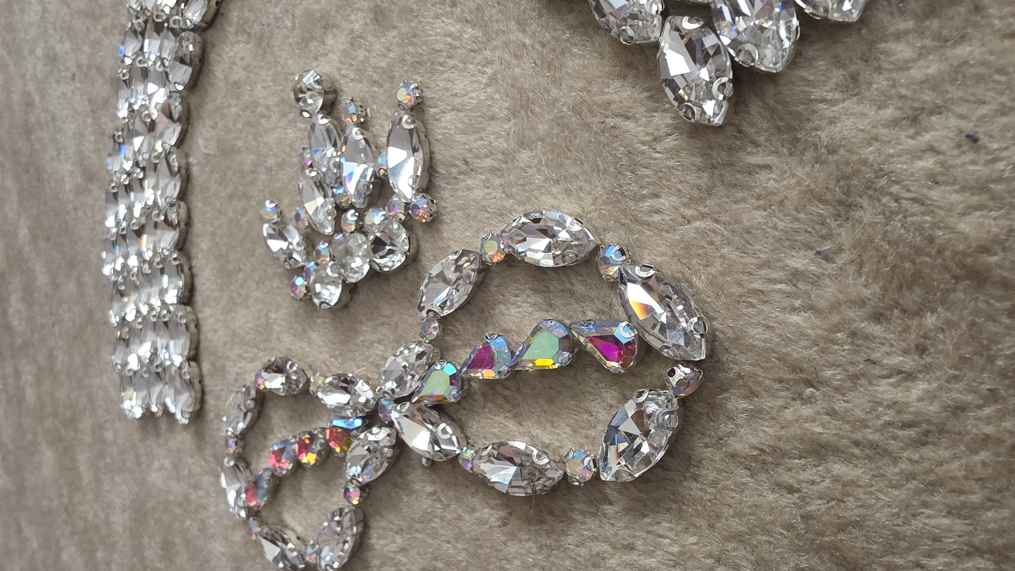 The image displays a close-up, high-angle view of various arrangements of sparkly, faceted rhinestones or crystals laid out on a textured, light brown or beige fabric surface, possibly a plush rug or faux fur. The gems are primarily clear and reflective, set in silver-toned metal casings, and many feature an iridescent "Aurora Borealis" (AB) coating, which creates vibrant flashes of green, pink, purple, and yellow as they catch the light.
On the left, a long, straight line of uniformly sized, oval-shaped clear gems is arranged vertically. Towards the middle and bottom, a flowing, almost circular pattern is formed by a sequence of larger oval clear gems interspersed with smaller, triangular or teardrop-shaped iridescent gems, showcasing their rainbow spectrum. Another cluster of mixed-shape gems, both clear and iridescent, is visible in the upper-left and central areas. A portion of a larger cluster of clear, oval gems appears in the top right corner.
The lighting is bright and even, accentuating the brilliance and prismatic qualities of the gems. The fabric background provides a soft, warm, and somewhat fuzzy texture. No activity is depicted; the items appear to be static, possibly awaiting use in crafting, fashion embellishment, or simply displayed. There are no people, specific location cues visible within the image itself, or any discernible text.