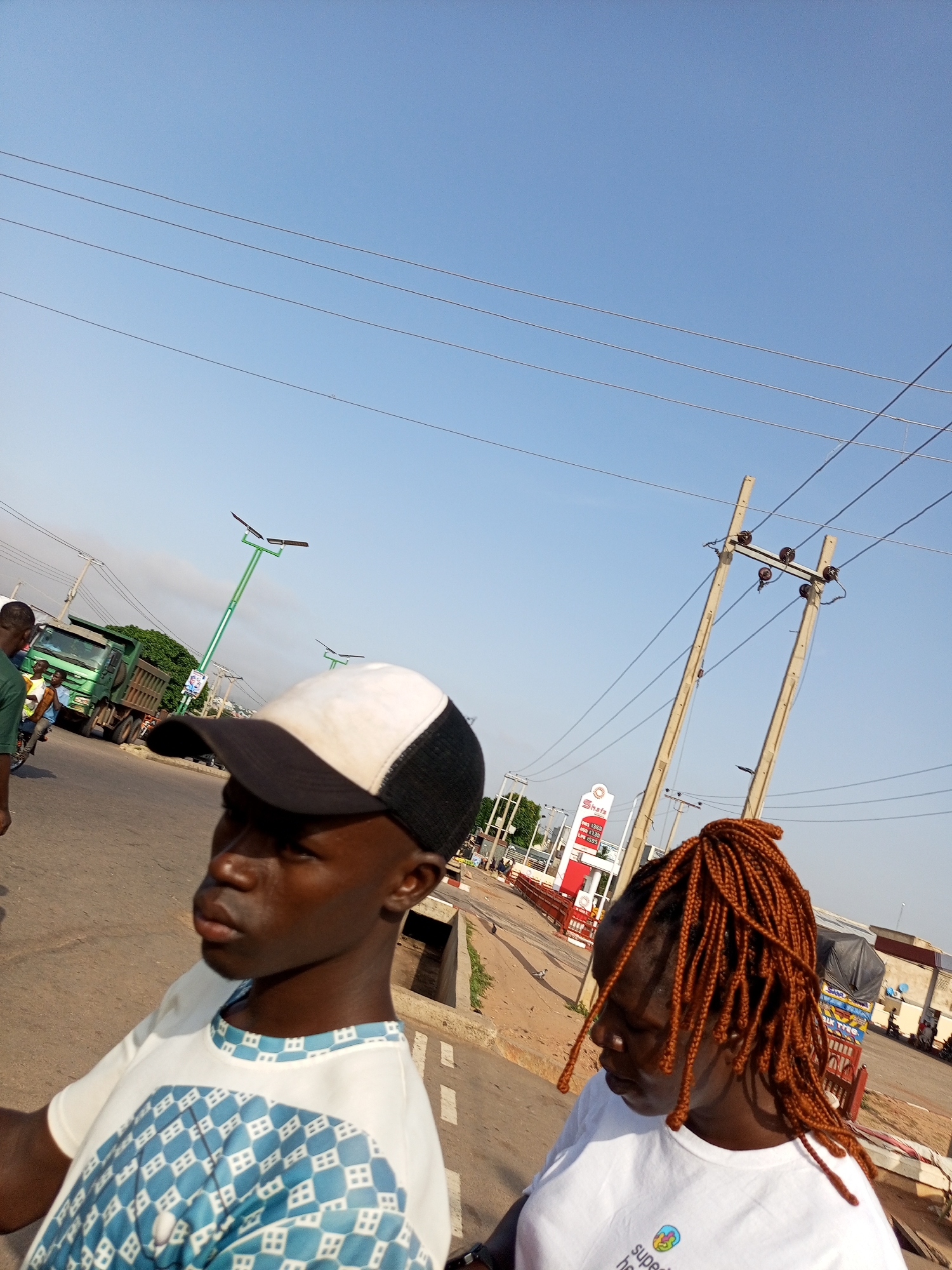 The image captures a street scene in Tumfure, Nigeria. In the foreground, two individuals are partially visible. The person on the left wears a white t-shirt with a blue and white patterned design and a black and white baseball cap. Their face is turned away from the camera, looking to the left. To the right, another person is seen from the shoulders up, wearing a white t-shirt. They have long, braided, reddish-brown hair and are looking downwards.
The background reveals a street with traffic and urban infrastructure. A green dump truck is parked on the left side of the road. Several streetlights with solar panels are visible, along with power lines and poles. A gas station with red and white signage, labeled "Shafa," is also present. The sky is clear and blue. The overall scene suggests a typical day in a Nigerian city.