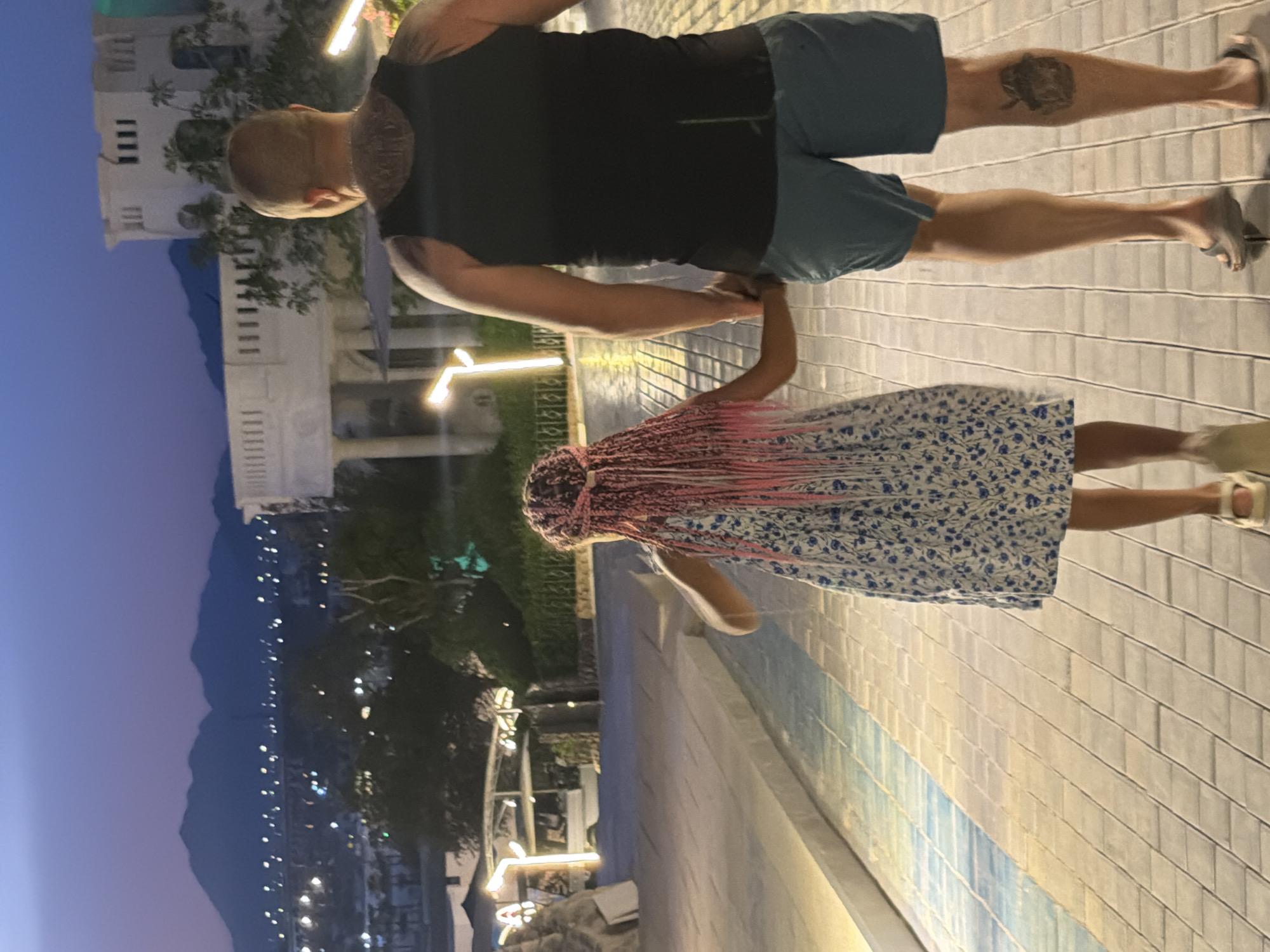A father and daughter are walking hand-in-hand on a tiled path in Sharm El Sheikh, Egypt, at dusk. The father, bald and wearing a black tank top and blue shorts, has tattoos on his back and left calf. The daughter, with pink braided hair, wears a blue and white floral dress. In the background, distant mountains are silhouetted against a purple sky, with city lights and modern white architecture visible. The path is illuminated by warm, horizontal light fixtures.