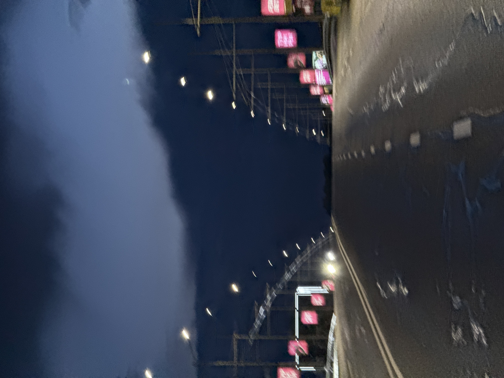 The image shows a dark, slightly blurry view of a road at dusk or night. The sky is a deep blue-gray with some wispy clouds. Along the sides of the road are poles with overhead wires, suggesting an urban or suburban environment. There are streetlights illuminating the scene, and some bright pink billboards are visible on the right side of the road. In the distance, there are headlights from an approaching vehicle. The road surface appears wet, possibly due to rain or recent cleaning. The scene is devoid of people. Without specific landmarks, it is impossible to determine the exact location within Minsk, Belarus.