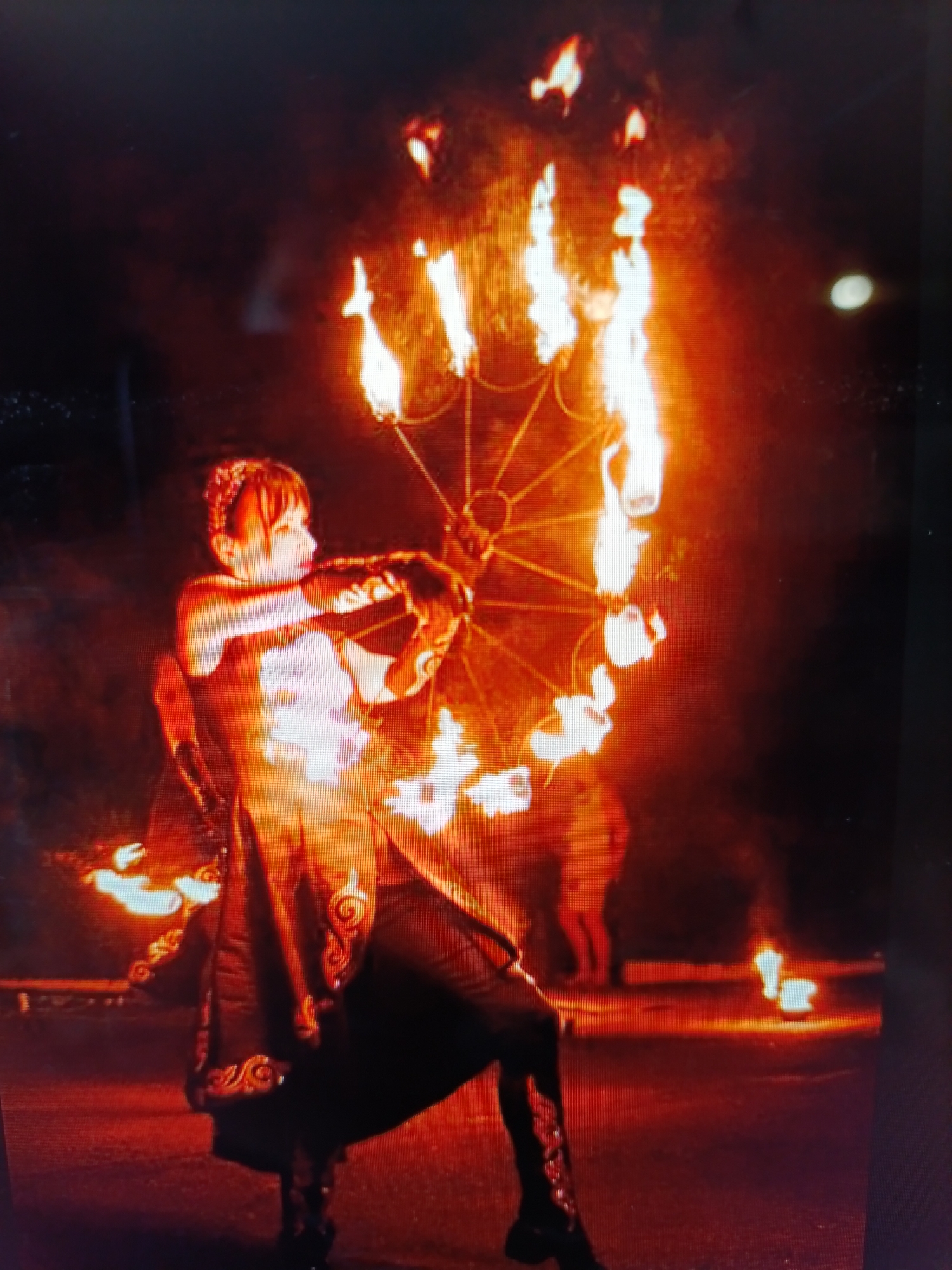 A performer is shown mid-performance in a dimly lit outdoor area. The performer, dressed in dark clothing with ornate trim, is holding a large, circular prop adorned with numerous flames. The prop is radiating heat and light, casting a warm glow on the performer and the surrounding environment. Smoke and embers are visible in the air, suggesting the intensity of the flames. In the background, other figures and small fires can be seen, indicating this is likely a public event or show. The ground appears to be paved, possibly a street or outdoor venue. The overall scene is dramatic and visually striking due to the contrast between the dark surroundings and the vibrant flames.