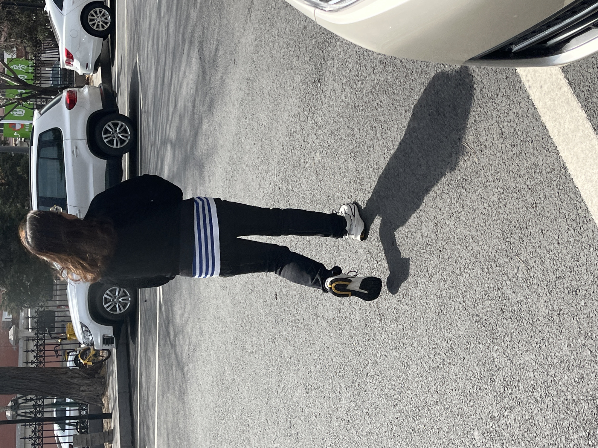 A pedestrian stands on an asphalt surface in a parking lot in Changchun City, China. The pedestrian is wearing a black jacket with blue and white stripes on the back and black pants. The pedestrian is also wearing running shoes that are black, white and gold. The parking lot is near a street with cars parked along the side.