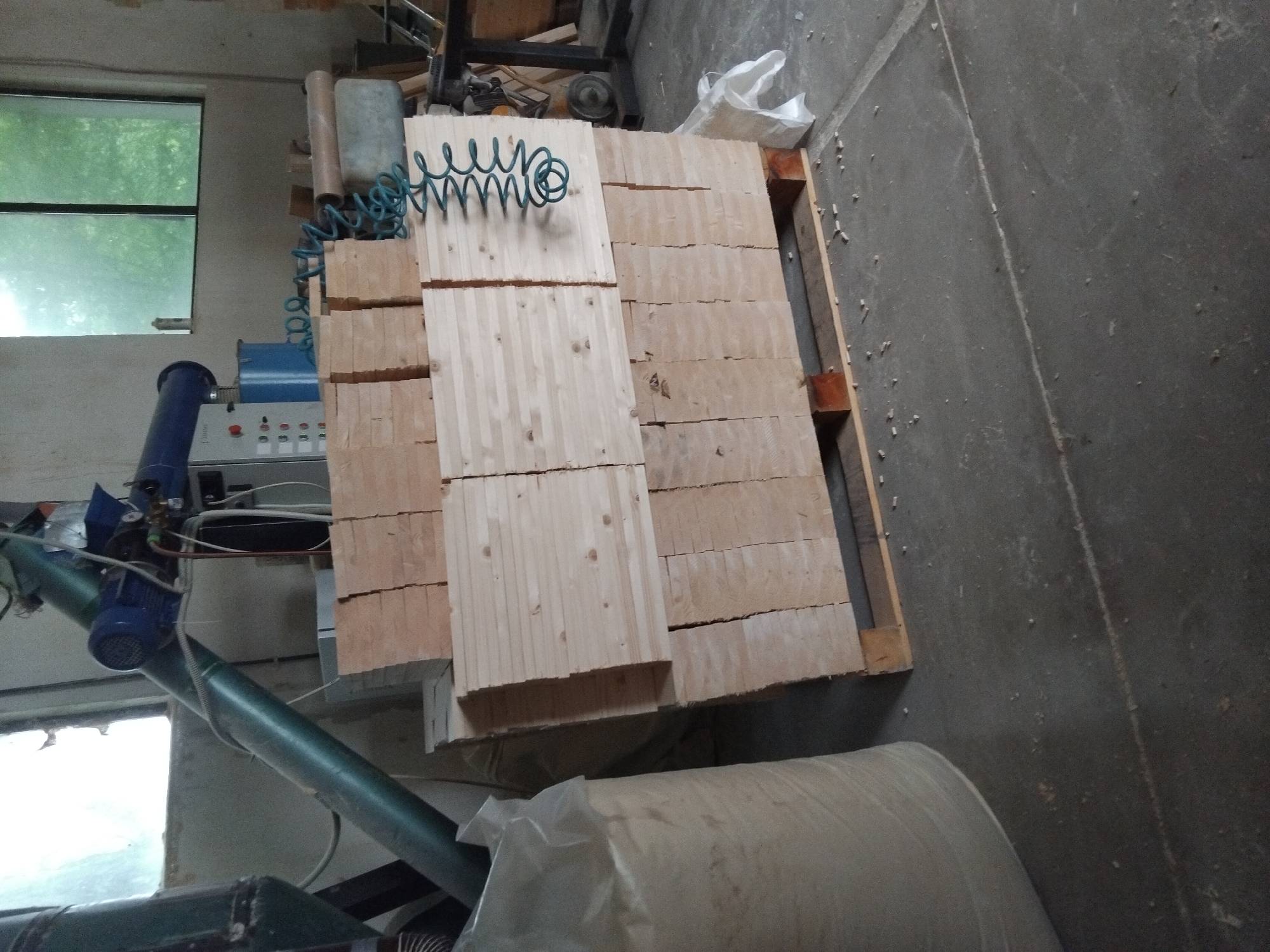 This image shows a pallet of stacked wooden planks inside a building, likely a workshop or industrial space. The planks are arranged in layers, with some appearing to be made of solid wood and others constructed from multiple smaller pieces joined together. A coiled blue air hose rests on top of the stack. To the left of the pallet, there's machinery, including a blue and grey piece of equipment with various controls. A large bag, partially obscured by a plastic cover, sits near the bottom right corner of the image. The location is not explicitly stated in the image.