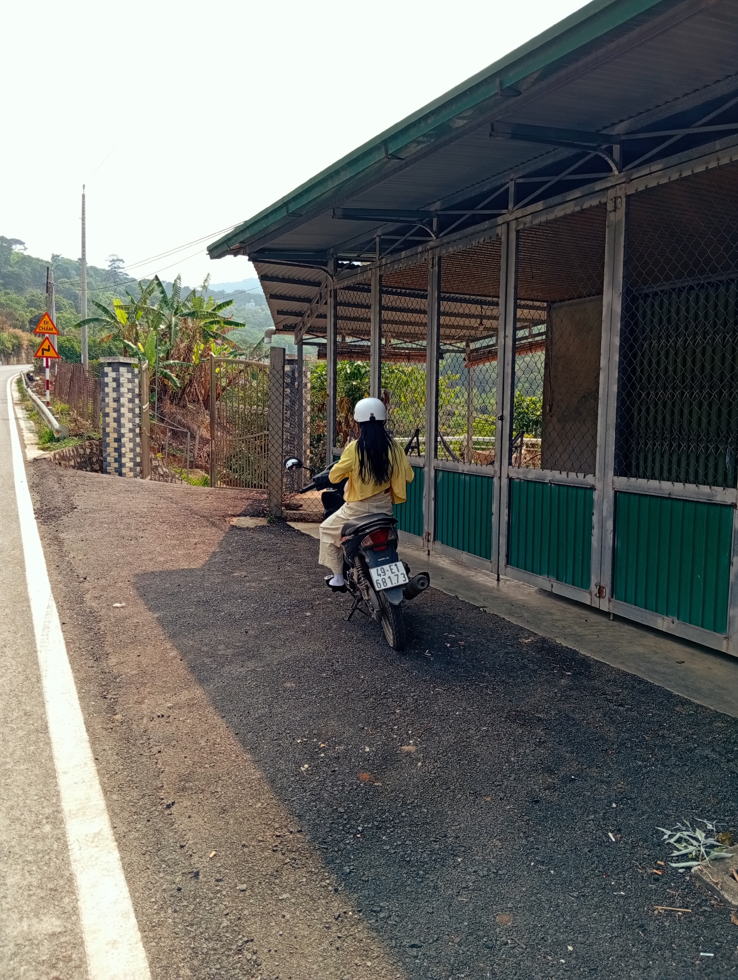 A person on a black scooter is depicted from behind, paused on the side of an asphalt road in a rural or semi-rural setting in D'Ran, Vietnam. The rider wears a yellow long-sleeved shirt, light-colored trousers, and a white helmet. The scooter's license plate displays "49-E1 681.73".
The road, marked with a distinct white edge line, curves gently uphill to the left. A prominent yellow triangular warning sign with the Vietnamese text "ĐI CHẬM" (meaning "Go Slow") is visible on the roadside, accompanied by another yellow warning sign indicating a double curve. Beyond a metal guardrail and a decorative brick and metal fence, a lush, vegetated hillside with banana trees rises.
To the right of the scooter is a long, low building, possibly a series of enclosures or storage units. It features a green corrugated metal roof supported by metal beams, and its front sections are composed of chain-link fencing above green corrugated metal panels. A simple metal gate leads into the property further along.
In the background, green mountains and hills stretch under a bright, clear sky, suggesting daytime conditions. Soft shadows are cast on the ground, indicating either morning or late afternoon sun. The image captures a serene moment of pause or travel in a scenic Vietnamese landscape.