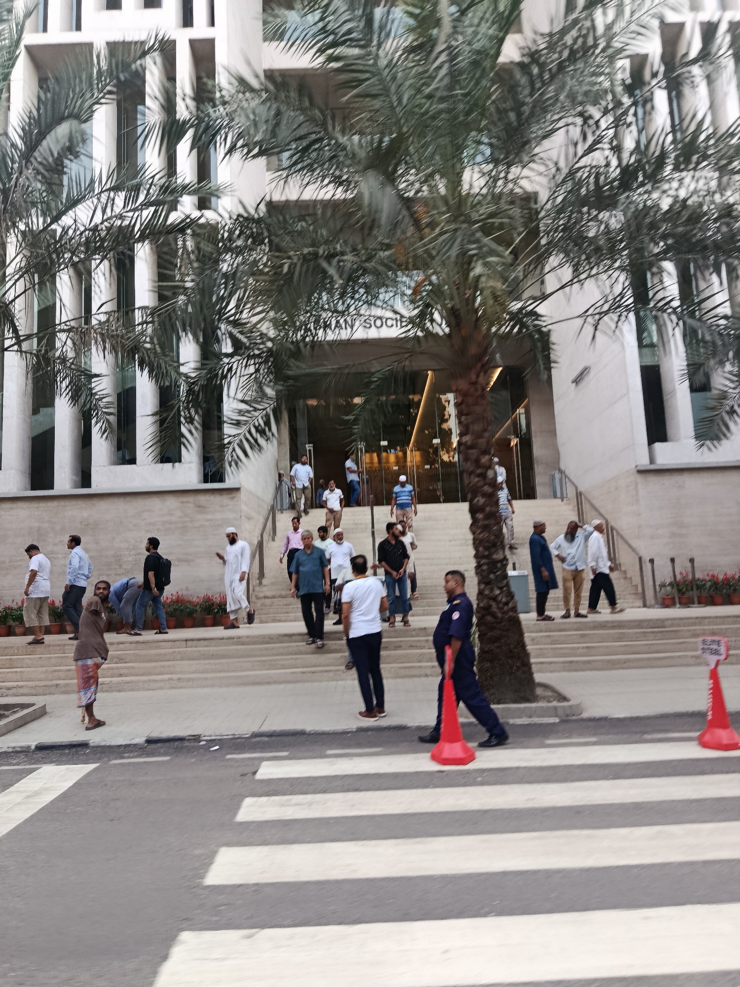 The image shows a group of people in front of a building in Dhaka, Bangladesh. The building has a sign that reads "HUMAN SOCIETY" above its entrance. People are walking both up and down the steps leading to the building. A palm tree obscures the entrance, with its trunk centered in front of the doorway. The people are dressed in a variety of clothing, with some wearing traditional Muslim attire. A security guard in a navy blue uniform walks across a zebra crossing in the foreground, passing an orange traffic cone. Another orange cone is visible on the far right of the crossing. Flower pots are arranged on the steps of the building. The architecture of the building is modern, featuring vertical rectangular windows. The sky is overcast.