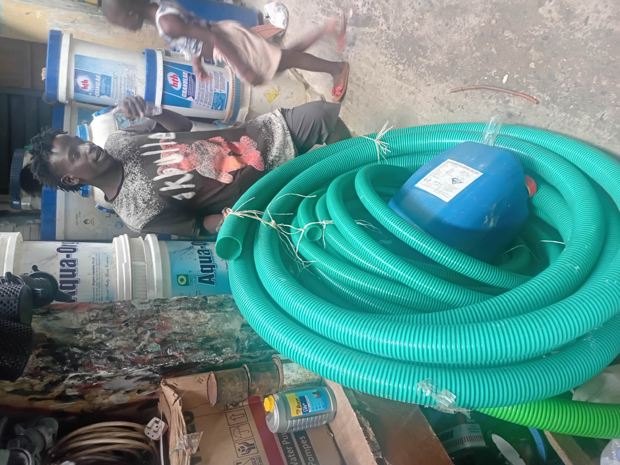 The image shows a young man standing in what appears to be a store or storage area, holding a bottle of water. He is wearing a dark t-shirt with a graphic print and dark pants. Behind him are stacks of large white plastic containers with blue lids, some labeled "hth GRANULAR" and "Aqua-Org". To his left, a large coil of bright green corrugated tubing is prominent in the foreground, partially obscuring a blue plastic container. A smaller grey plastic container with a yellow cap and a label that reads "FIXIT" is also visible. In the background, there is a wall with colorful paint splatters and some hanging items. A child, mostly out of focus, is visible to the right of the man, walking away. The ground is concrete. The overall environment suggests an industrial or commercial setting, possibly a hardware store or a place selling agricultural or cleaning supplies. The scene is lit by artificial light, casting some shadows.