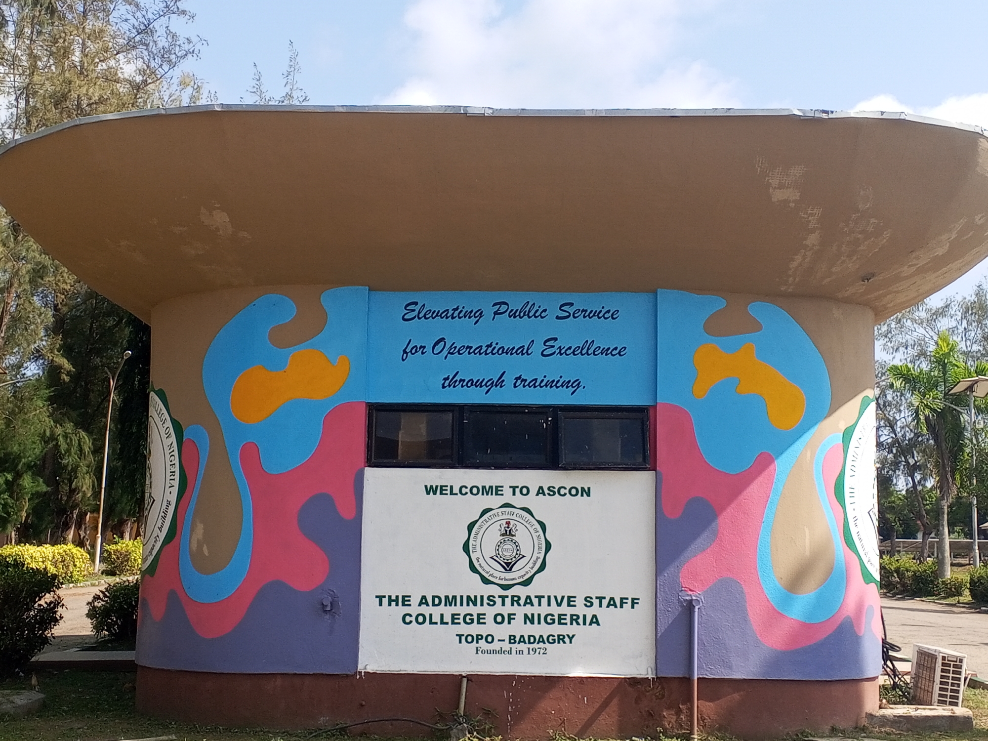 The image shows a low, rounded, painted structure with an overhanging roof, located outdoors. The structure bears the inscription: "Elevating Public Service for Operational Excellence through training." Below this, a sign reads "WELCOME TO ASCON" above a logo, followed by "THE ADMINISTRATIVE STAFF COLLEGE OF NIGERIA, TOPO-BADAGRY, Founded in 1972". Abstract blue, pink, purple, tan, and yellow designs adorn the sides of the structure. There are windows above the welcome sign. Vegetation, including trees and bushes, surrounds the structure. An air conditioning unit sits to the right of the structure. The sky is visible in the background.