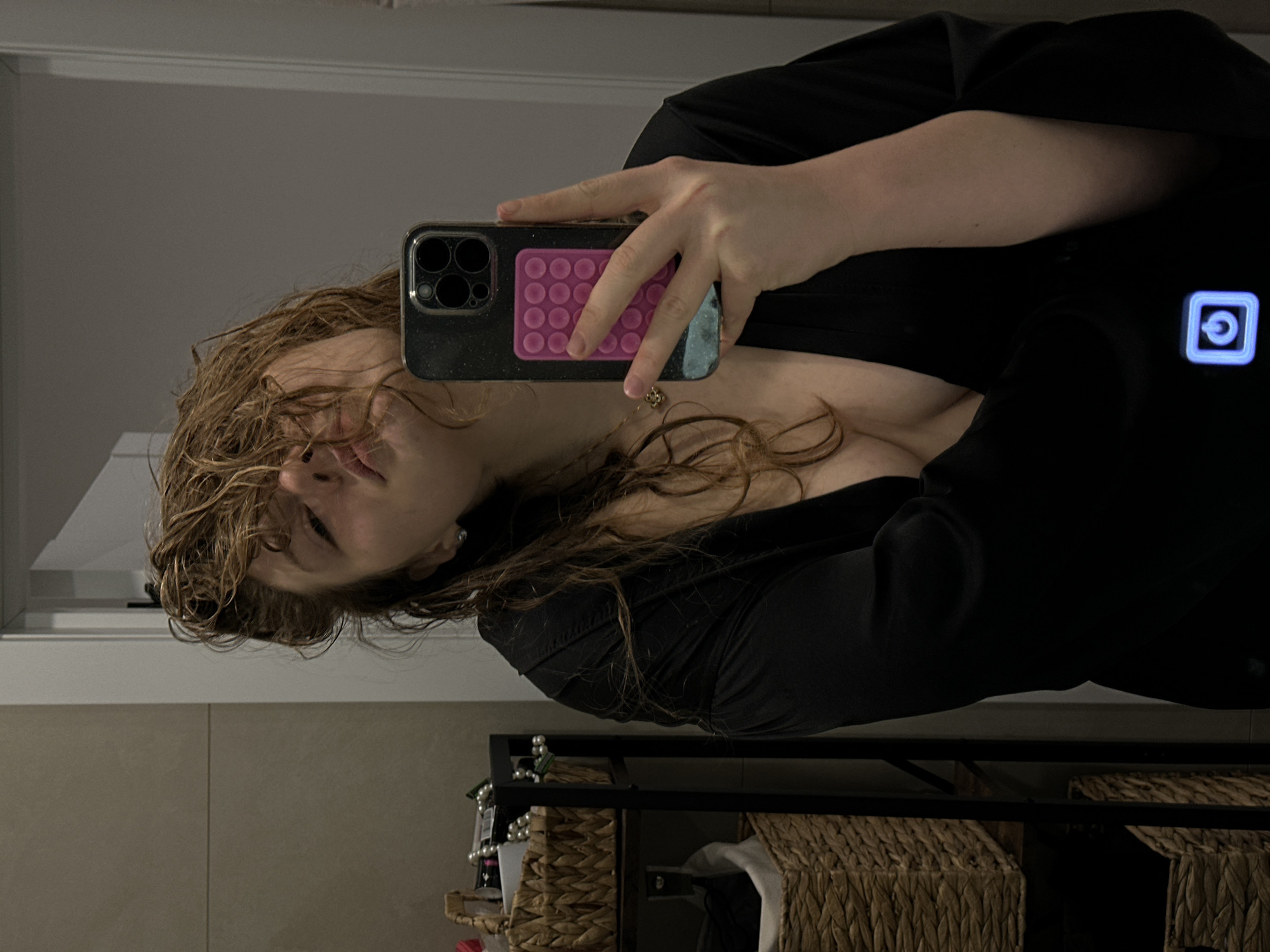 A woman is taking a selfie in a bathroom or dressing area. She is holding a black iPhone with a pink pop-it case. She is wearing a black garment with a blue illuminated power button. Her hair is curly and partially covers her face. Behind her, there is a countertop with beige tiles and a black metal shelf unit with woven baskets. There is no indication in the image that this is in the city of Studenets, Russia.
