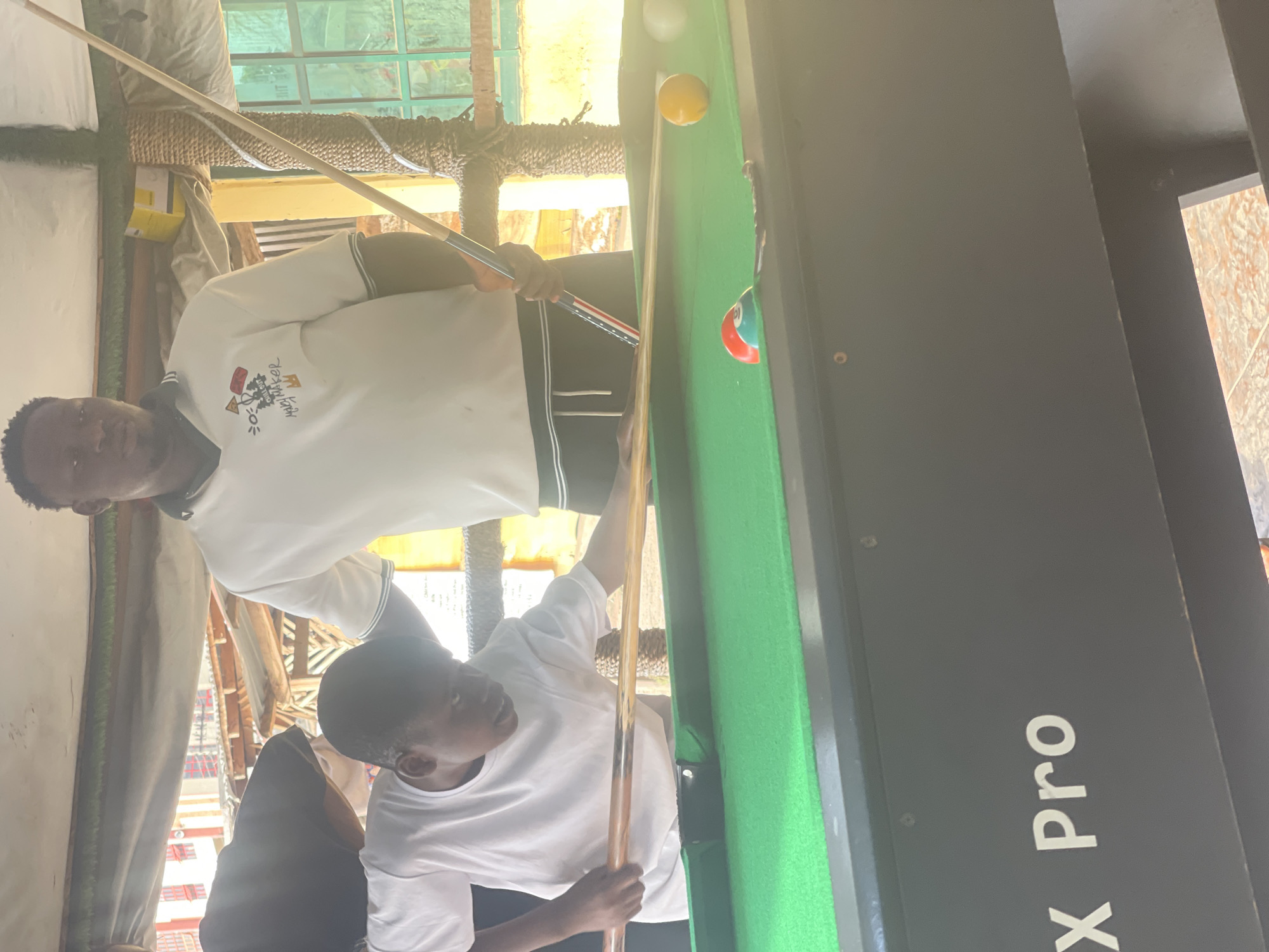 Two young Black men are playing pool indoors. The man on the left wears a white long-sleeved polo shirt and is holding a pool cue over the shoulder of the man on the right, who is in a white t-shirt and is aiming the cue towards the pool table. The pool table is green with white and yellow balls. "X Pro" is printed on the side of the table. The location is likely indoors in Lokoja, Nigeria.