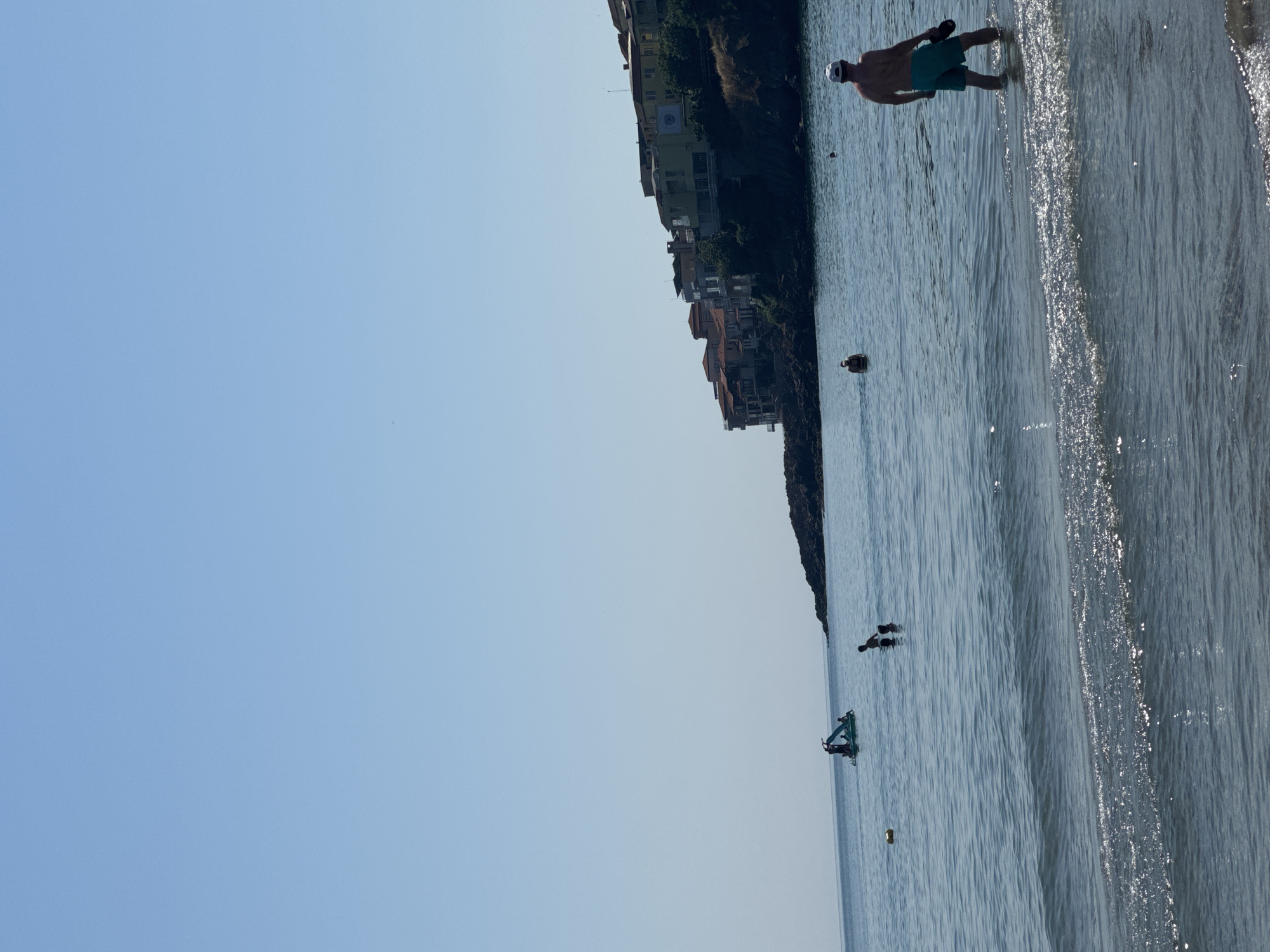 The image depicts a sunlit day at a beach in Sozopol, Bulgaria, likely along the coast near the old town, given the presence of cliffside buildings in the background. The water is calm with gentle waves lapping at the shore. In the foreground, a man wearing teal shorts and a white cap stands in the shallow water, facing away from the camera and holding a dark object, possibly a bag, in his right hand. Further out in the sea, several other people are visible. To the left, a small, green boat-like watercraft with several individuals on it is present. Near the center, two people are standing in the water. In the middle ground, a yellow buoy floats. The background features a rocky cliff densely populated with buildings, many of which have red-tiled roofs, characteristic of Sozopol's historic architecture. The sky is clear and blue, indicating fair weather.