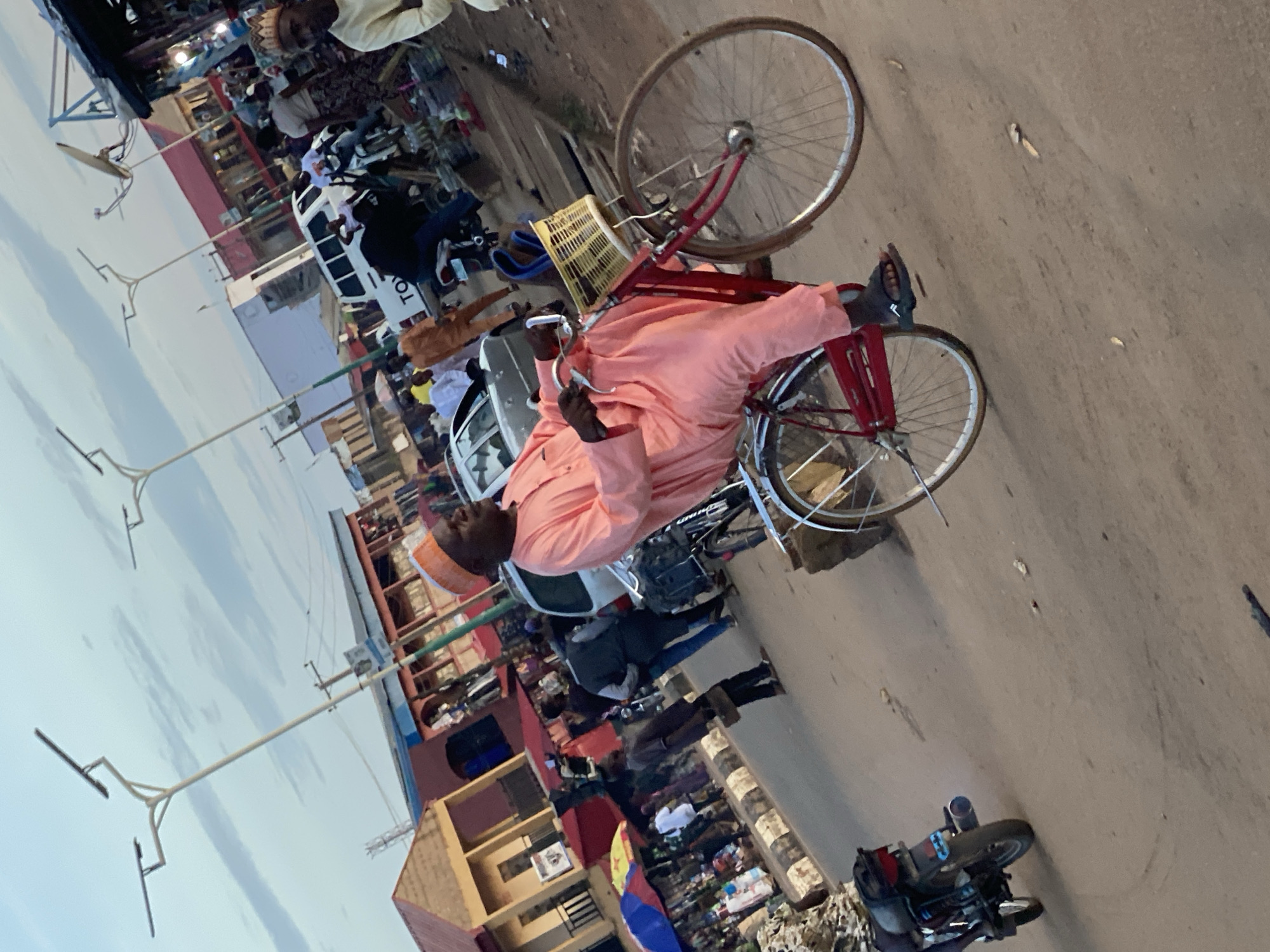 This image depicts a bustling street scene in Kafanchan B, Nigeria, observed during daylight hours under a partly cloudy sky. The primary subject is an adult male, of apparent middle age, riding a red bicycle equipped with silver fenders and a light-colored woven basket attached to the handlebars. He is dressed in a peach-colored traditional tunic and a matching cap, with dark sandals on his feet. He maintains a relaxed posture, hands on the handlebars, and a smile or laugh evident on his face as he looks towards the right.
The road surface is unpaved and appears to be dirt or compacted earth, light brown in color with some scattered debris. In the mid-ground, a white Toyota pickup truck is present, along with multiple pedestrians and other individuals operating motorcycles. To the far left, a black motorcycle is partially visible. The background features various commercial structures, including single and two-story buildings, some with red roofs or blue accents, and multiple market stalls identifiable by colorful umbrellas (blue and red are discernable). Numerous individuals are seen walking and congregating throughout the background, indicating active commerce. Modern streetlights, characterized by multiple horizontal arms with integrated solar panels, ascend towards the sky. On the far right, a man in light-colored attire and a patterned cap is partially visible, and a satellite dish is mounted on a distant rooftop.