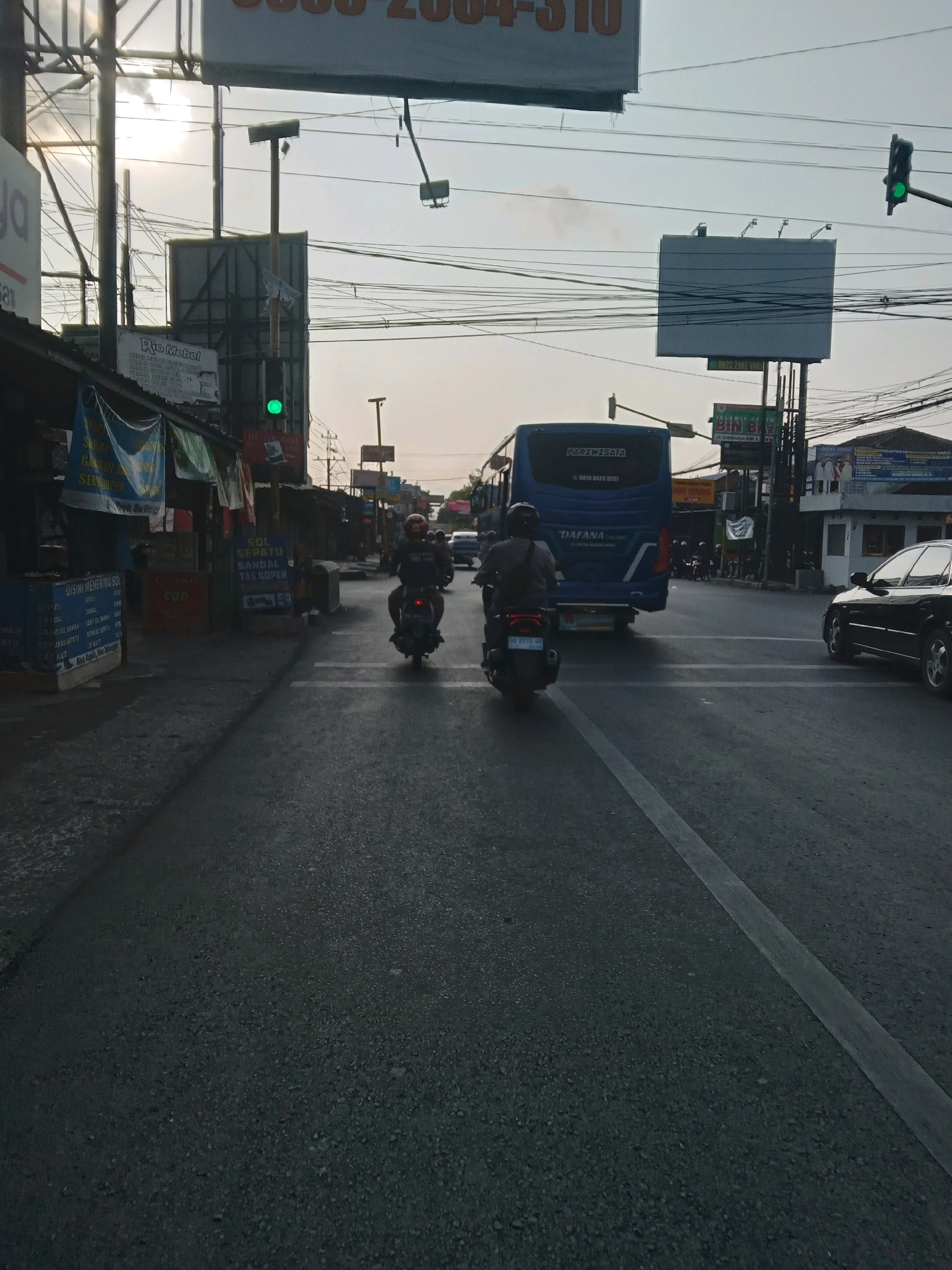 The image depicts a street scene in Sitimulyo, Indonesia, likely during daylight hours with overcast skies. A blue bus with "PARIWISATA" and "DAFANA" visible on its rear is in the center lane of the road. Two individuals on motorcycles are also present, one in front of the bus and one to the left. The motorcycle in front of the bus has a rider wearing a red helmet and a dark jacket, while the motorcycle to the left has a rider wearing a grey jacket and a dark helmet.
On the left side of the road, there are several shops or stalls with banners displaying various text, including "RIO MEBEL" and "DIBUTUHKAN PENGAMBILAN GAMBAR". A green traffic light is visible on a pole. To the right of the bus, a black car is parked or moving in the right lane. The road surface is asphalt, marked with white lines. Utility poles and numerous overhead wires crisscross the scene. Billboards are also visible in the background. The overall impression is of a busy, urban street environment.
