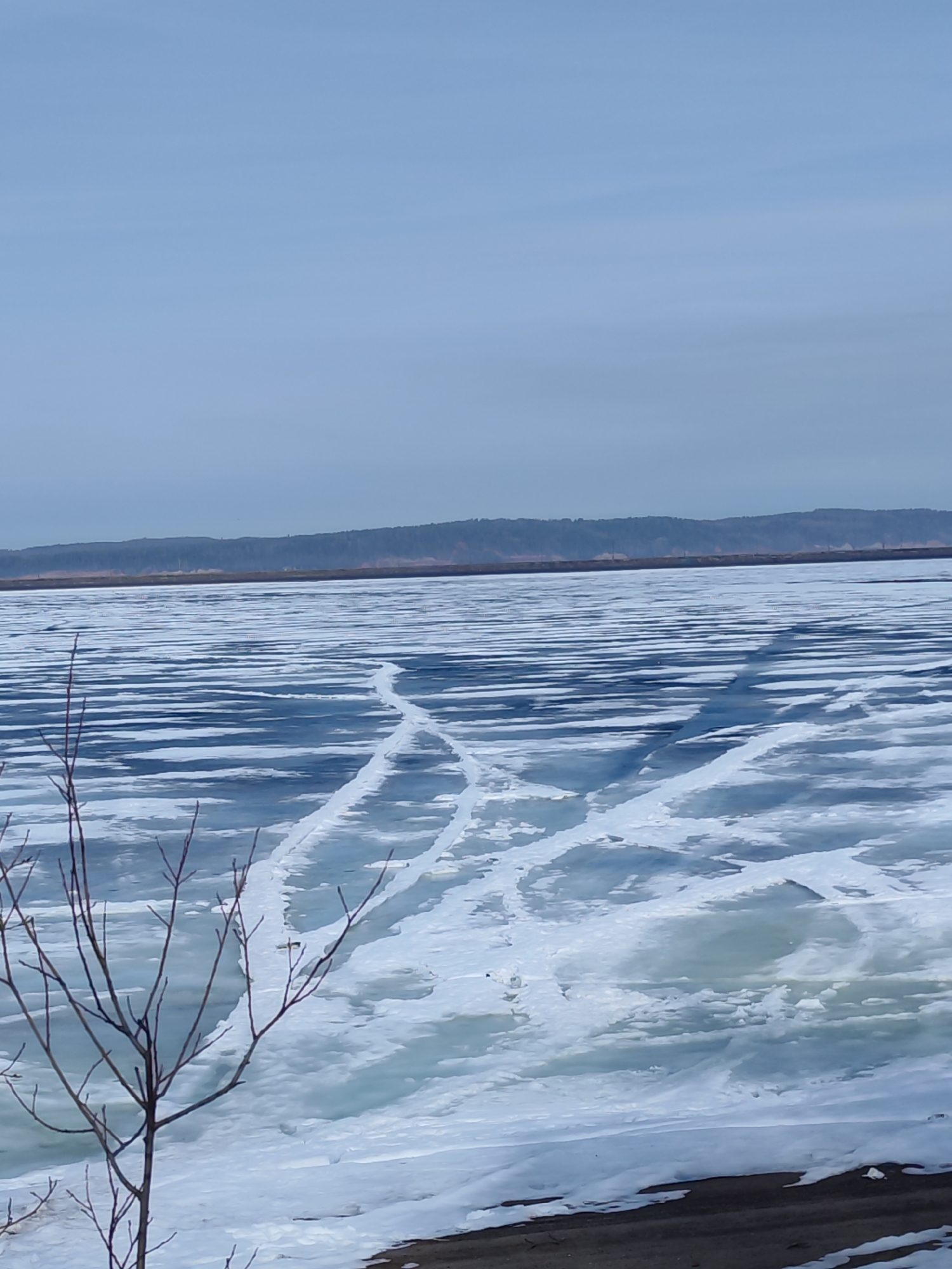 The image depicts a winter scene on the Kama River in Chaikovsky, Russia. The river is largely frozen over, with cracks and varying ice formations visible. Linear tracks run across the ice surface, suggesting a vehicle or repeated foot traffic. In the foreground, a bare tree stands with its branches reaching upwards. The riverbanks are covered in snow. In the distance, the horizon line shows forested hills. The sky is overcast, creating a muted light across the scene.