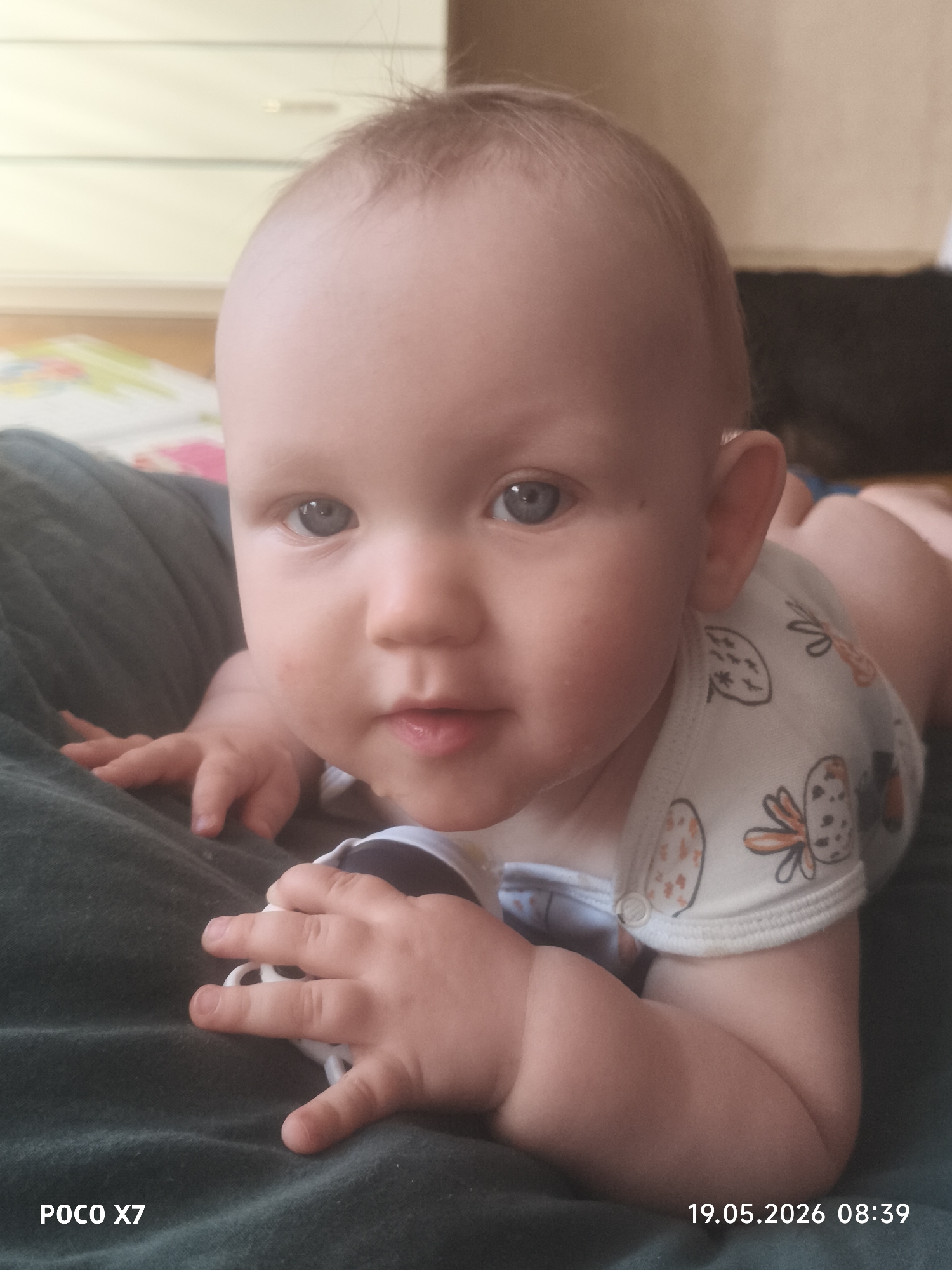 A close-up shot depicts a baby lying prone, supported by a green, possibly fabric, surface. The baby has fair skin, light-colored hair, and blue eyes. The baby is wearing a white onesie with cartoon-like drawings of fruit and butterflies. The baby’s face is the main focus, with a slightly open mouth and visible saliva. The baby's hands are visible in the foreground, resting on the green surface.
In the background, out of focus, is a light-colored piece of furniture and a patterned object or fabric.
The image has been stamped with the date "19.05.2026 08:39" and the device "POCO X7".
There is no city or specific location mentioned in the image.