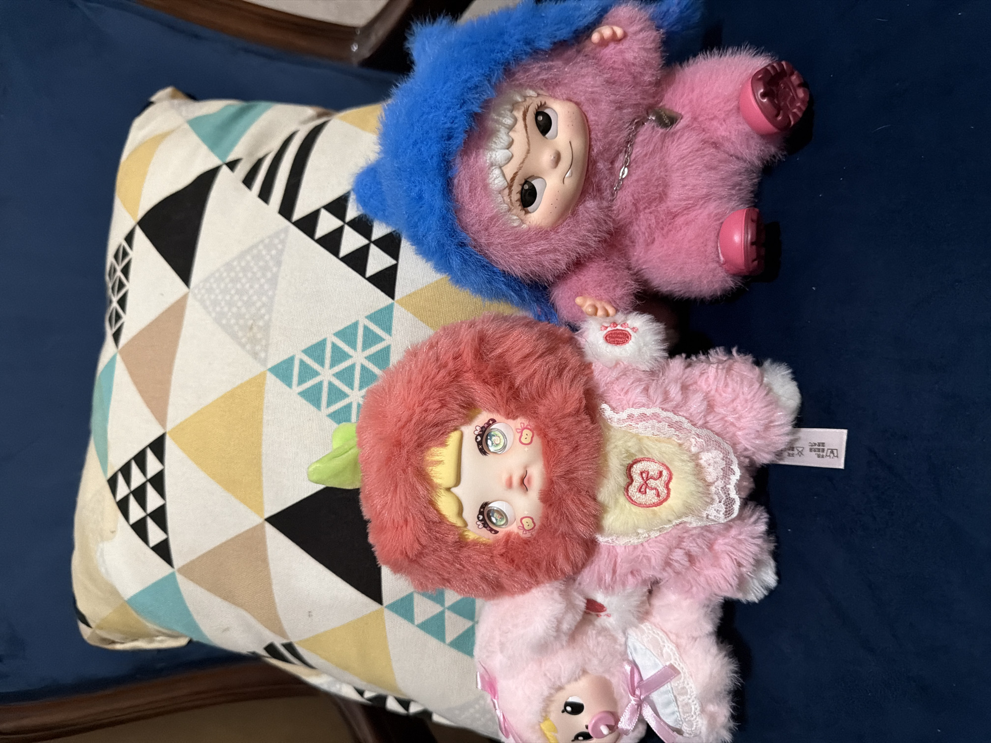 Two pink dolls, one with a blue hood and one with a red hood, are lying on a patterned pillow on a blue surface. The scene is located in East Jakarta, Indonesia.