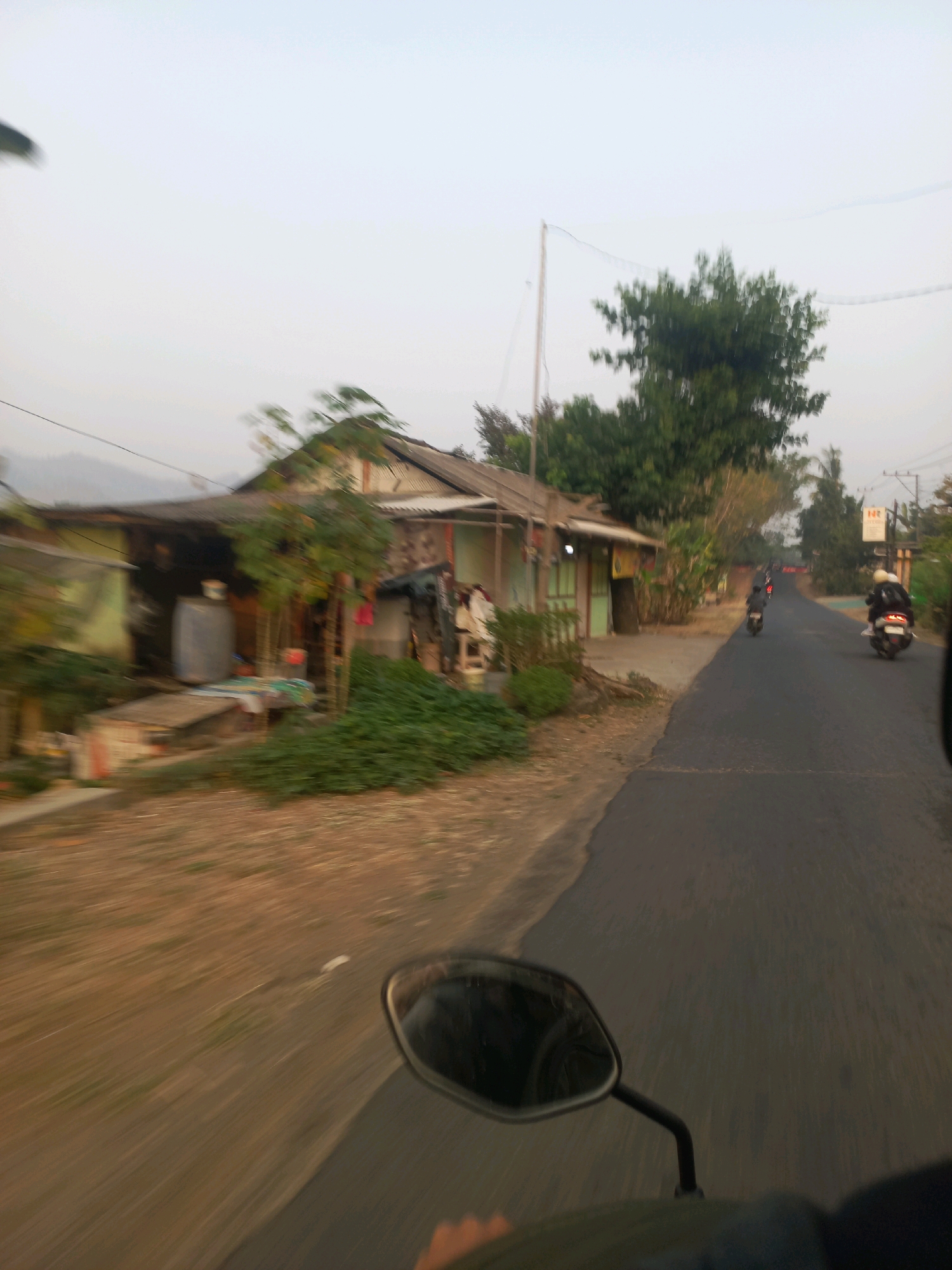The image depicts a street scene in Ngarjosari, Indonesia, likely during daylight hours with overcast skies. The perspective is from a moving vehicle, possibly a motorcycle, as indicated by the visible rearview mirror in the foreground. To the left of the road, there are several modest buildings, appearing to be homes or small businesses, with corrugated metal roofs and simple facades. Vegetation, including bushes and trees, is present around the buildings and along the roadside. A paved road runs through the scene, with several motorcycles visible in motion. One motorcycle with two riders is in the foreground on the right side of the road. Further down the road, other motorcycles are also seen, suggesting traffic. The environment appears to be rural or semi-urban, characterized by the simple architecture and natural elements. The overall impression is one of a typical everyday scene on a road in this Indonesian location.