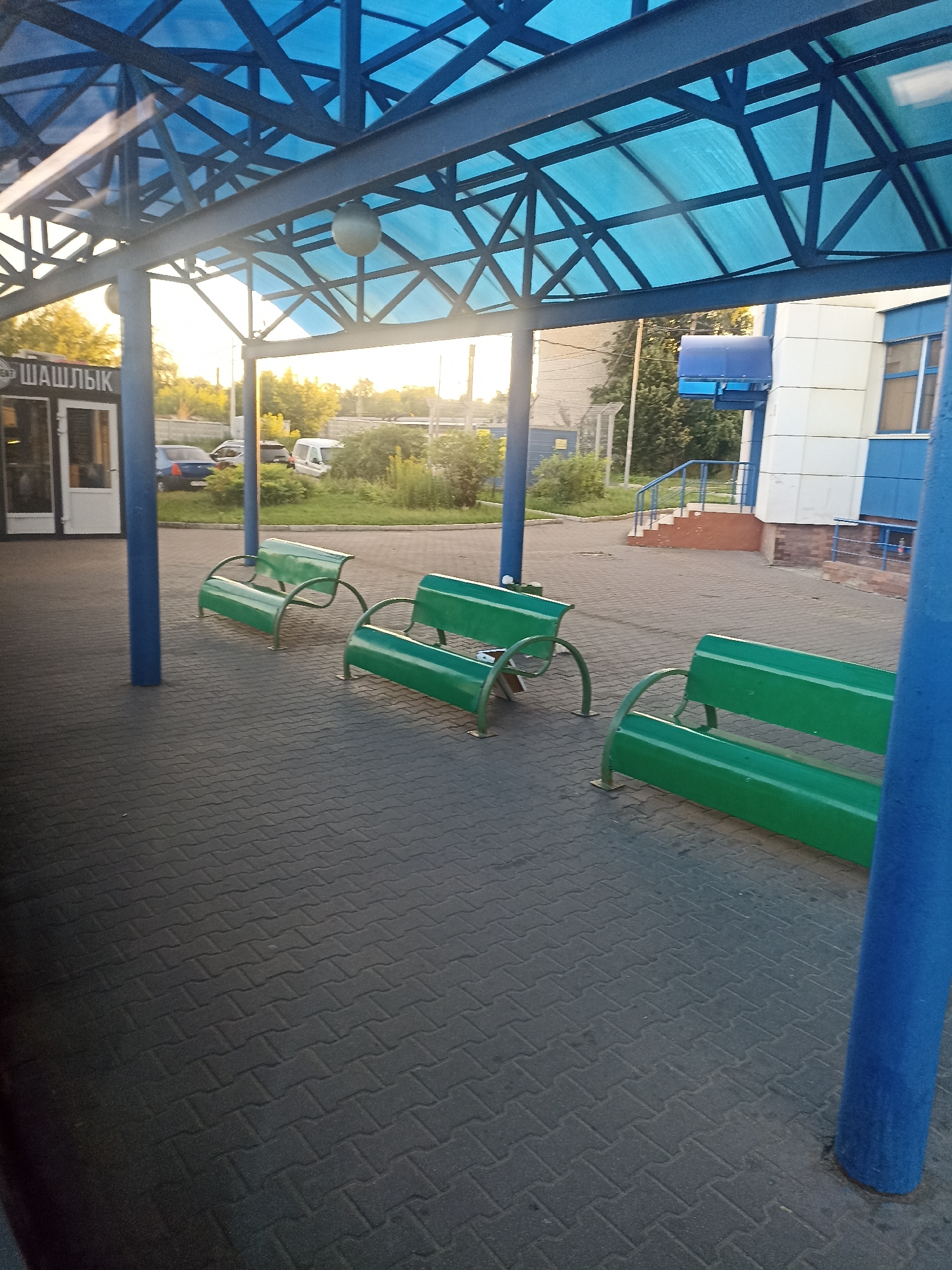 A sunny day at a bus stop in Chekhov, Russia. The scene is captured from a low angle, looking outwards from under a blue canopy. In the foreground, a dark grey brick pavement is visible. Three green benches are arranged in a row on the pavement. Behind the benches, a building with white and blue panels is partially visible, with a set of stairs leading up to it. The background shows trees and a road with a few cars parked. The sky is bright and clear, suggesting good weather.