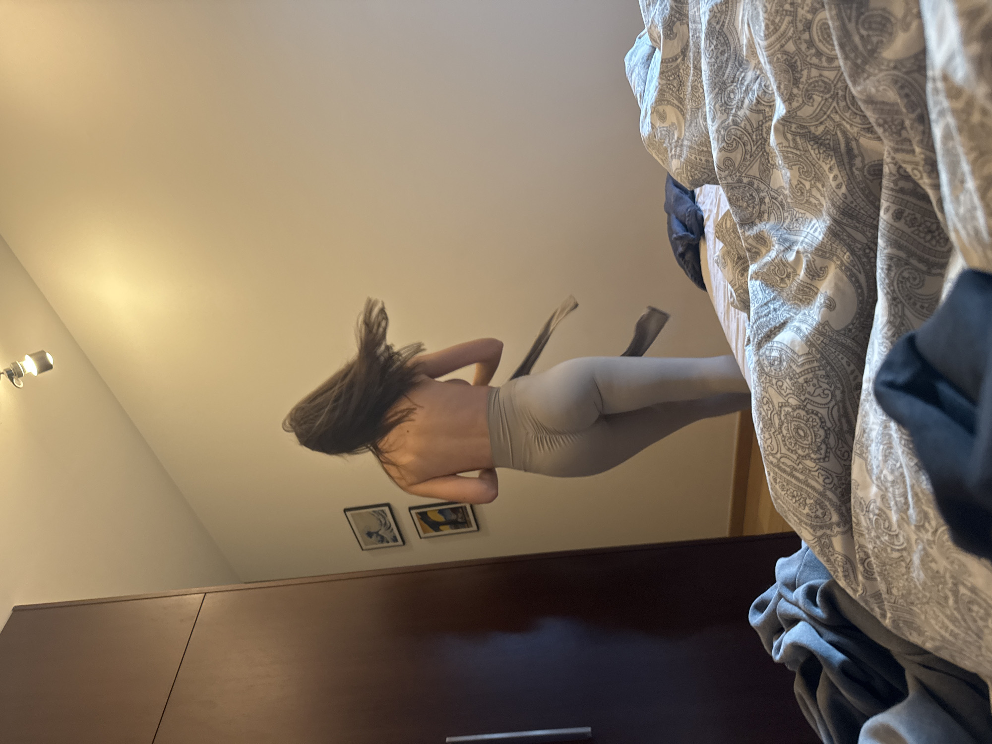 A low-angle, wide shot captures a partially nude woman in light-colored leggings. She is oriented diagonally in the frame, with her head towards the upper left and her legs towards the upper right. Her back is bare, and her hair is loose. Two black objects are suspended from her head. Behind her, on the wall, are two framed pictures. Below the pictures is a large brown wardrobe. A bed with a patterned comforter is visible in the upper right corner. The location is presumed to be an indoor setting within Warsaw, Poland.