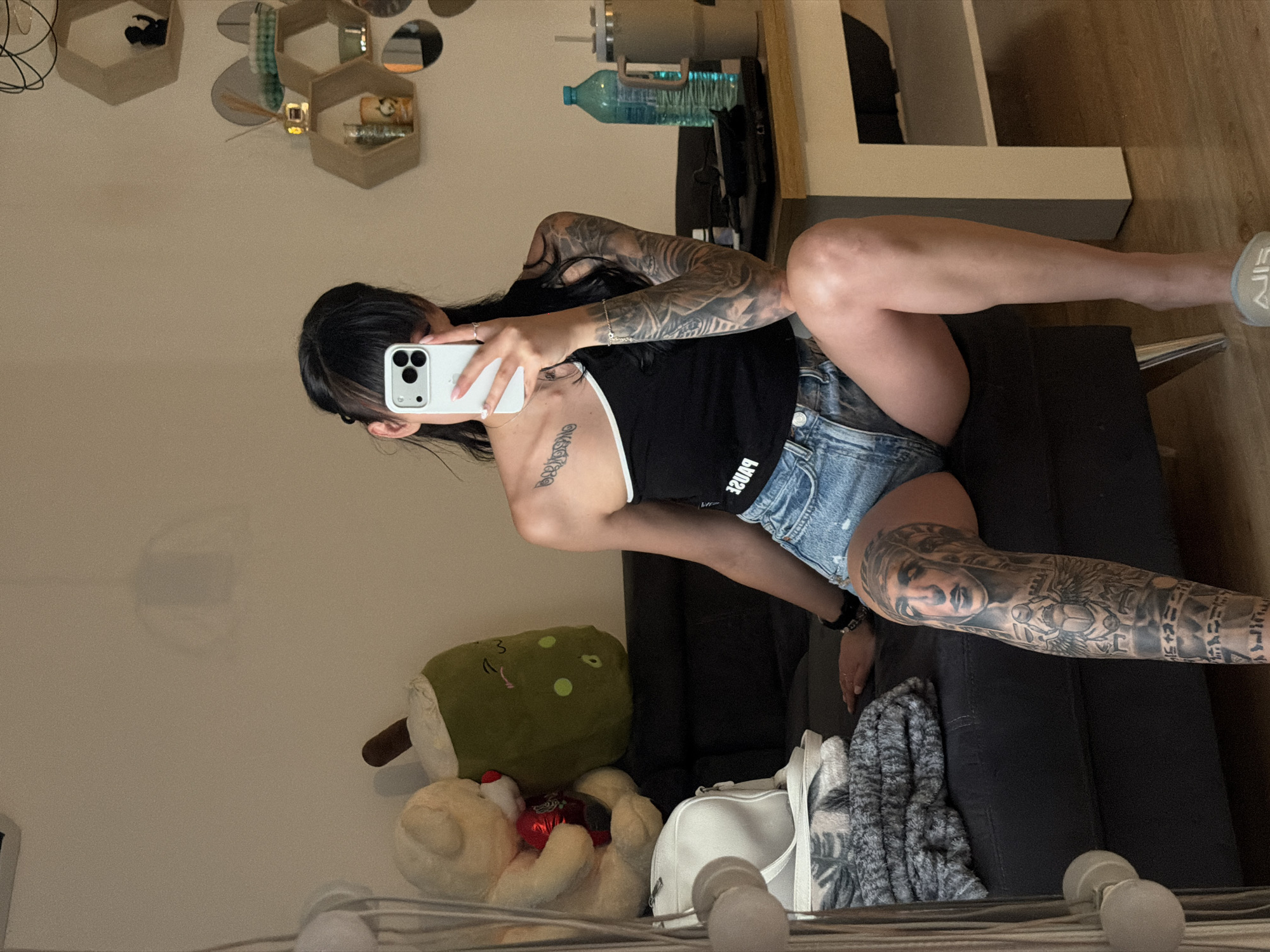 The image shows a woman with dark hair and bangs, holding a smartphone and taking a mirror selfie. She is wearing a black one-shoulder top with white trim and denim shorts. Her left arm and left leg are covered in tattoos. She is sitting on a dark sofa. To her left, there is a white handbag and a stack of folded gray blankets. Behind the sofa, there are two plush toys: a teddy bear holding a heart-shaped object and a green bubble tea cup. The background features a cream-colored wall with hexagonal shelves displaying decorative items and a water bottle on a wooden table. The scene appears to be indoors, likely in a residential setting. No specific location in Plovdiv, Bulgaria, is indicated by any visual cues in the image.