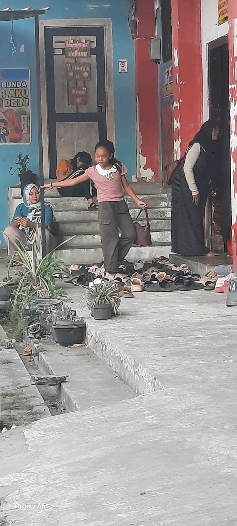 The image depicts an outdoor scene in front of a building, possibly a community center or school, in Selopuro, Indonesia. A young girl wearing a pink shirt and grey pants is standing on a concrete staircase, holding a brown purse. To her left, an adult woman wearing a blue headscarf and grey pants is seated on the same staircase. Behind them, another person is seated further up the stairs, wearing an orange shirt and black pants. A large pile of various sandals and shoes is scattered on the ground at the base of the stairs. The building has a blue facade on the left with signage and a red facade on the right. Several potted plants are visible on the left side of the staircase. The environment appears to be a public space, with a focus on the activity on the stairs.