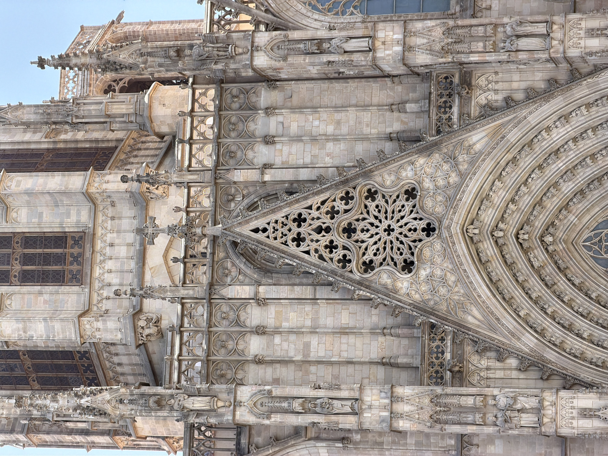 The image captures a detailed view of the Gothic facade of the Barcelona Cathedral, officially known as the Cathedral of the Holy Cross and Saint Eulalia, located in the Gothic Quarter of Barcelona, Spain. The perspective is frontal, focusing on the intricate architectural elements of the upper section of the cathedral.
The facade is constructed from light-colored stone, with a dominant Gothic architectural style characterized by pointed arches, ribbed vaults, and elaborate tracery. The central part of the image is dominated by a large, triangular gable above the main portal, filled with a complex rose window featuring quatrefoil and trefoil patterns. This gable is flanked by vertical buttresses and ornate spires.
Below the gable, a series of layered arches form the entrance, each decorated with numerous small sculptures or gargoyles. Niches are carved into the stone, housing statues of religious figures, some of whom are depicted in prayer or holding religious symbols. Figures are visible standing in niches on either side of the main entrance, and higher up, on decorative ledges.
The stone walls are textured with individual blocks, and decorative stringcourses and cornices add further architectural detail. Above the main gable and rose window, a balustrade with delicate lattice work runs horizontally, with more spires and finials extending upwards. Tall, narrow windows with Gothic tracery are visible in the background, suggesting the interior structure of the cathedral. The sky above is a clear, light blue, indicating a bright, sunny day.
There are no discernible people actively engaged in any actions within the frame. The focus is entirely on the static, monumental architecture of the cathedral. The scene is static and presents a close-up view of the rich ornamentation and craftsmanship of this historic building.