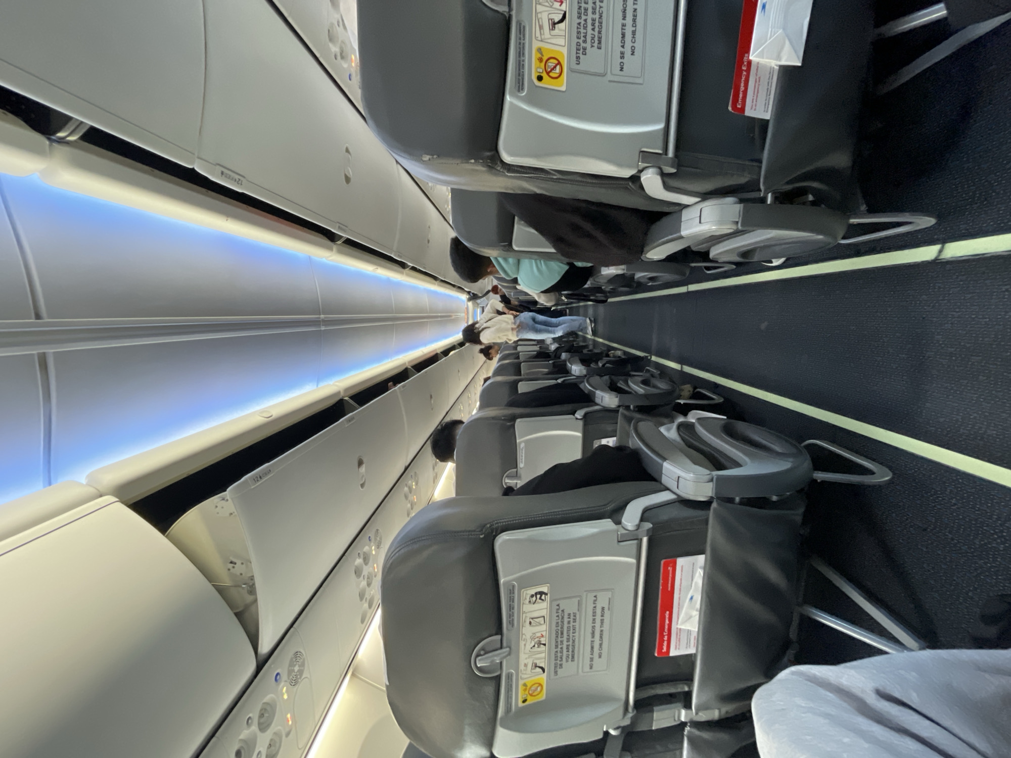 The original image captures the interior of an airplane cabin, rotated 90 degrees clockwise, presenting a view down a central aisle. Rows of dark grey passenger seats with black fabric backs line both sides, some with occupants and others empty. The perspective is from an upward angle, making the seats appear to hang sideways.
On the left, white overhead compartments feature a prominent blue LED strip light that illuminates the cabin. On the right, the seats have folded tray tables and safety information cards tucked into seat pockets. Passengers are visible further down the aisle, some seated and others appearing to be standing or moving. Their faces are not clearly discernible.
Notable details include the bright, artificial blue lighting along the ceiling, contrasting with the generally dim cabin. Emergency exit signs and information are prominently displayed. Visible text from the seatback in the foreground reads, "USTED ESTA SENTADO EN LA FILA DE SALIDA DE EMERGENCIA" / "YOU ARE SEATED IN AN EMERGENCY EXIT SEAT" and "NO SE ADMITE NIÑOS EN ESTA FILA" / "NO CHILDREN THIS ROW". A red sign indicates "Emergency Exits" / "Salida de Emergencia". Labels with the number "12" are visible on the overhead bins, likely indicating row numbers. The scene depicts a typical moment inside an aircraft, possibly during boarding, deplaning, or while inflight. The location context provided for the flight is Ushuaia, Argentina.