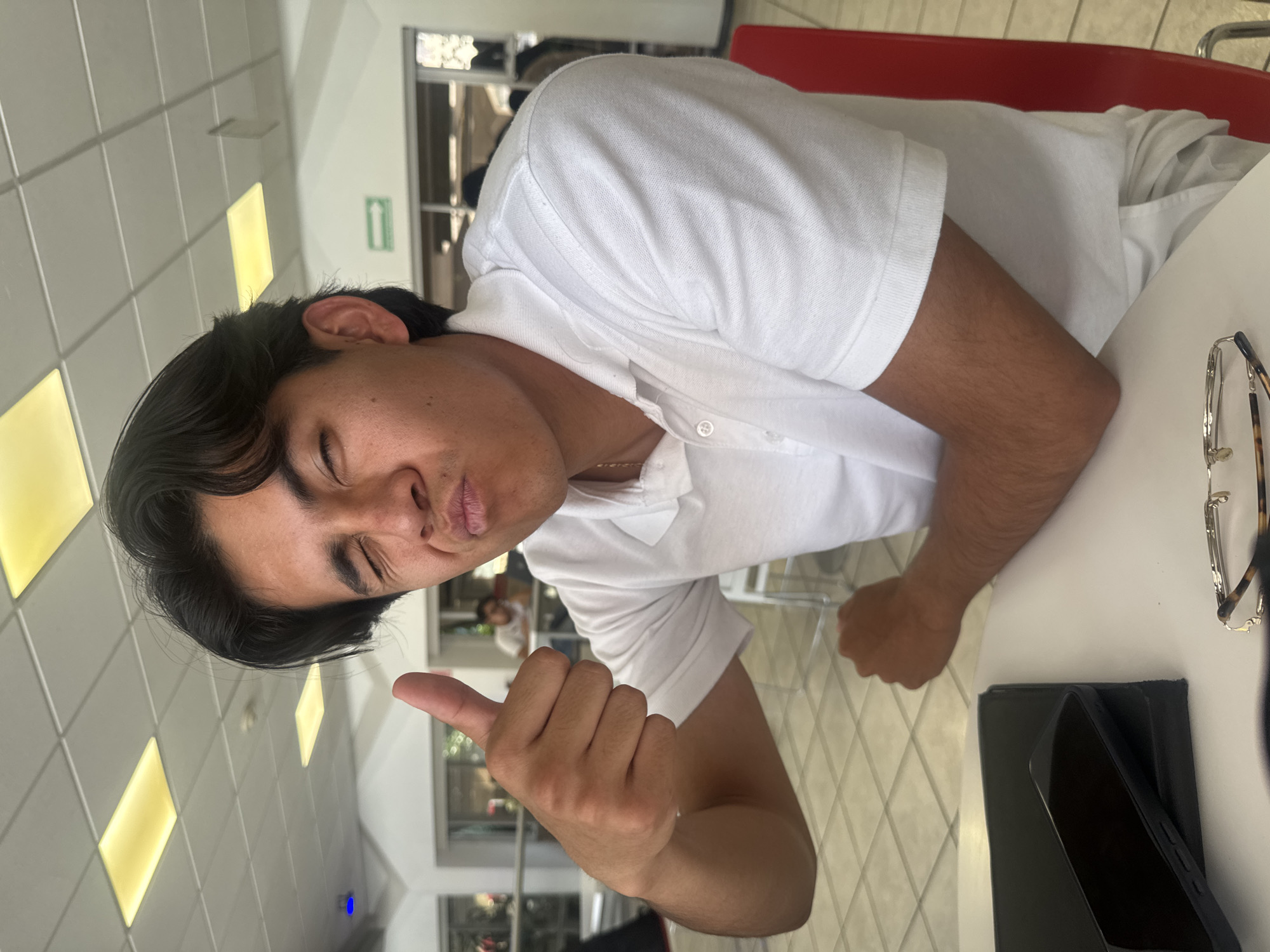 A person with dark hair is sitting at a table. They are wearing a white collared shirt and giving a thumbs up with their right hand. A pair of glasses and a phone are on the table. The location appears to be inside a building with windows and a tile ceiling. An exit sign is visible. The location could be in Puebla, Mexico.