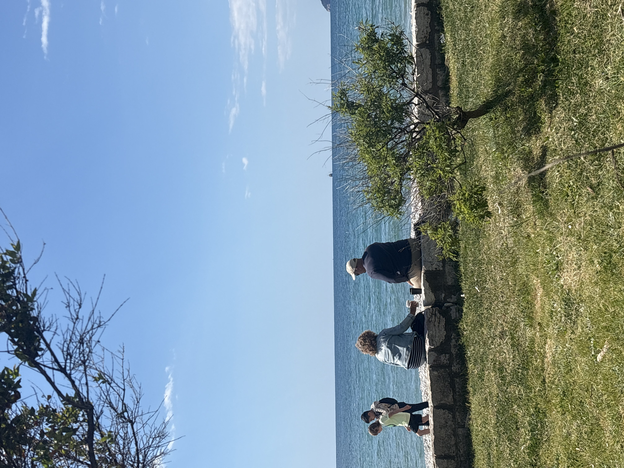 The image shows a group of people on a stone wall overlooking the Adriatic Sea. An adult man with a baseball cap is sitting on the wall on the right side of the image, holding a cup. A woman is sitting to his left on the wall, facing the sea. Two children are standing on the ground level beside the woman and interacting. The background shows the blue sea and the sky with some clouds. The area adjacent to the wall is covered in green grass and a tree is growing near the wall. This scene takes place on the seaside of Bar, Montenegro.