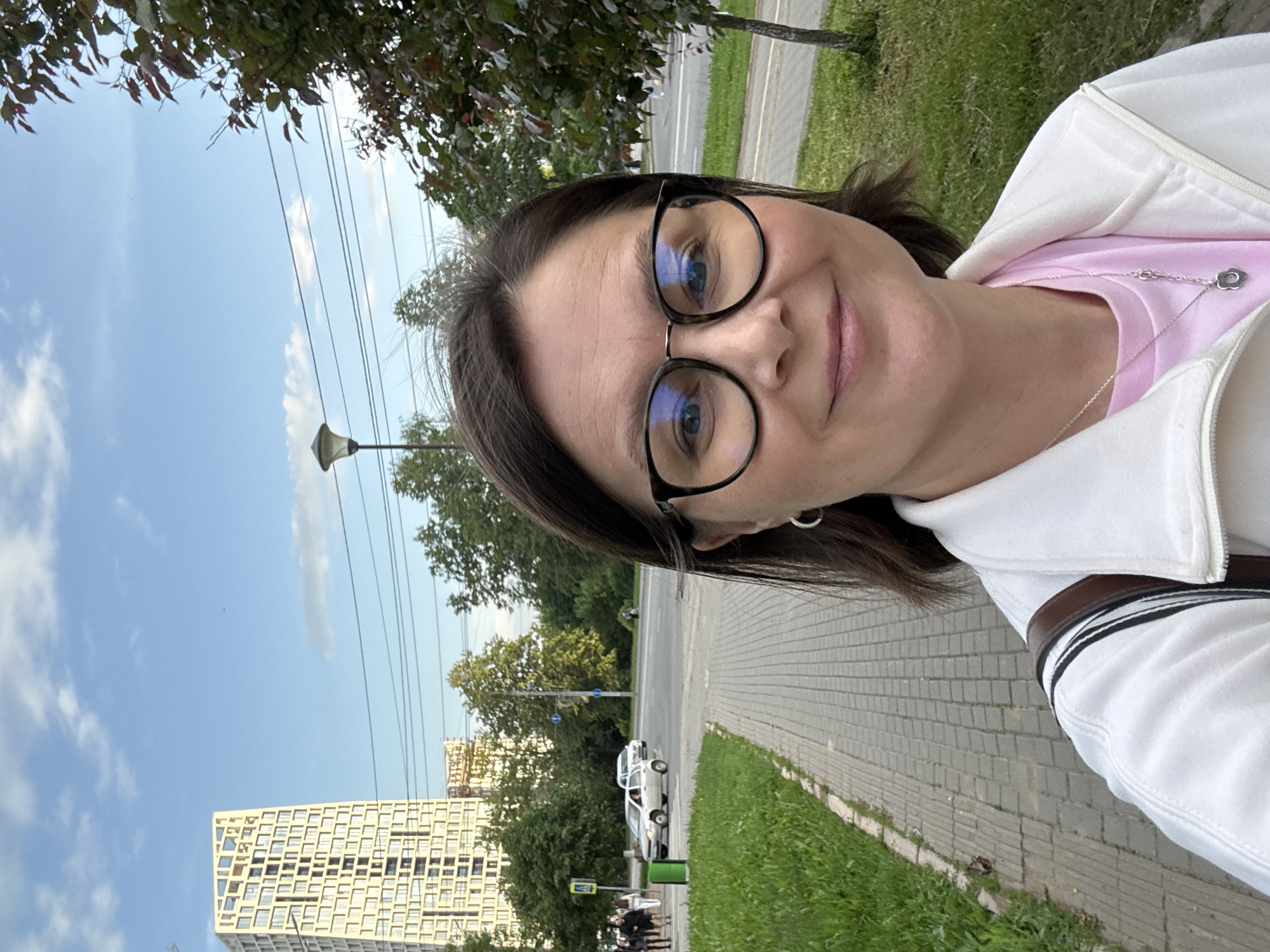 A woman, wearing glasses and a white zip-up jacket over a pink shirt, takes a selfie outdoors. She is smiling. A silver necklace with a flower pendant is visible. The background shows a street with cars, trees, a sidewalk, a tall building, and a blue sky with some clouds. Power lines are also visible. The image appears to be taken during the day, with good lighting and clear weather. The overall mood is casual and pleasant. The tall building in the background has a distinctive geometric facade.