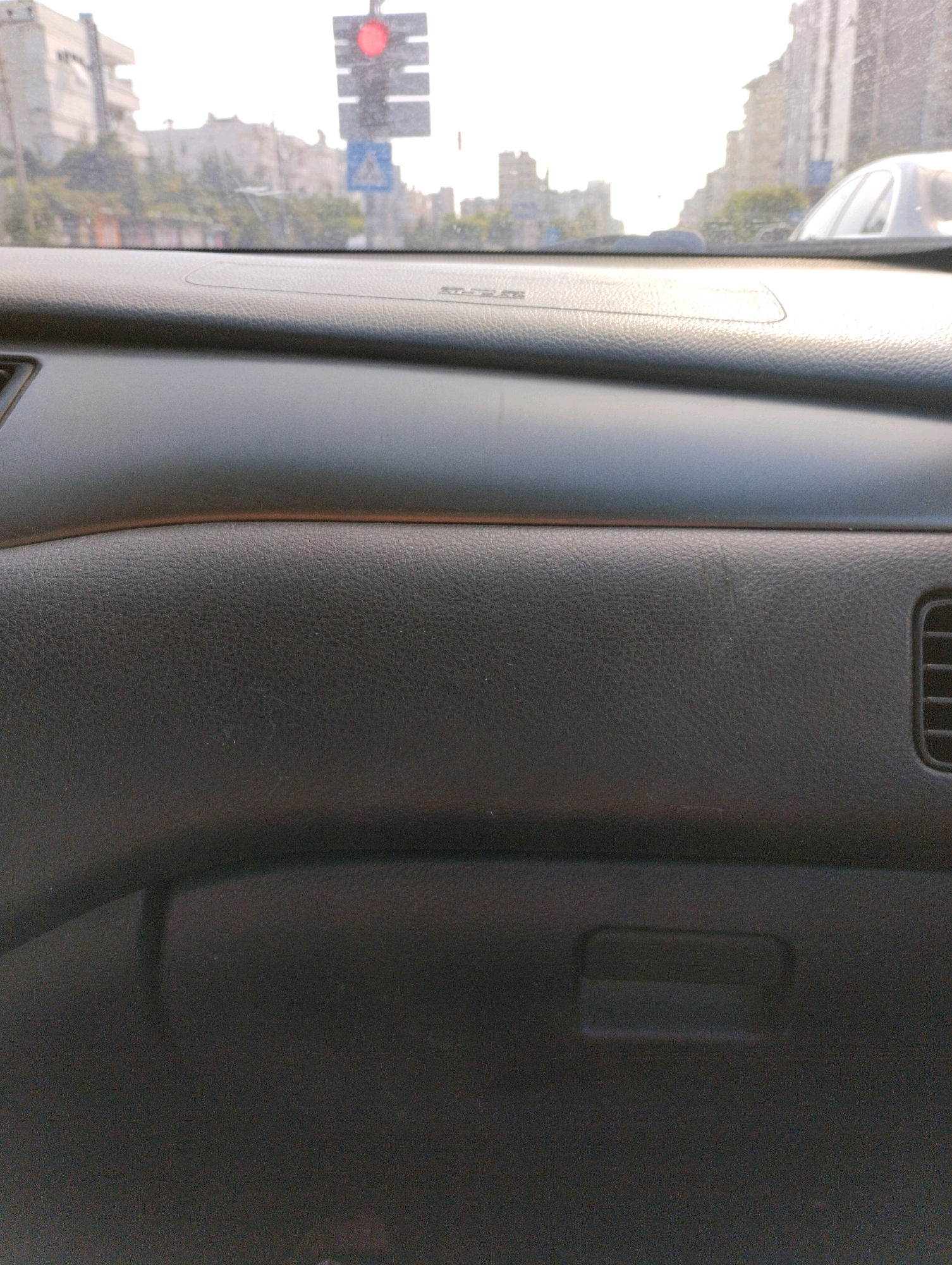 The image is a close-up view of a car's dashboard and windshield, taken from the interior of the vehicle. Through the windshield, a blurry cityscape is visible, characterized by buildings and a traffic light showing red. The dashboard is dark gray and textured, with a curved surface in the upper portion and a flatter section below, which appears to be the glove compartment. A vent is visible on the right side of the dashboard. The scene suggests the car is stationary, possibly at a traffic signal, in the city of Mezitli, Turkey.