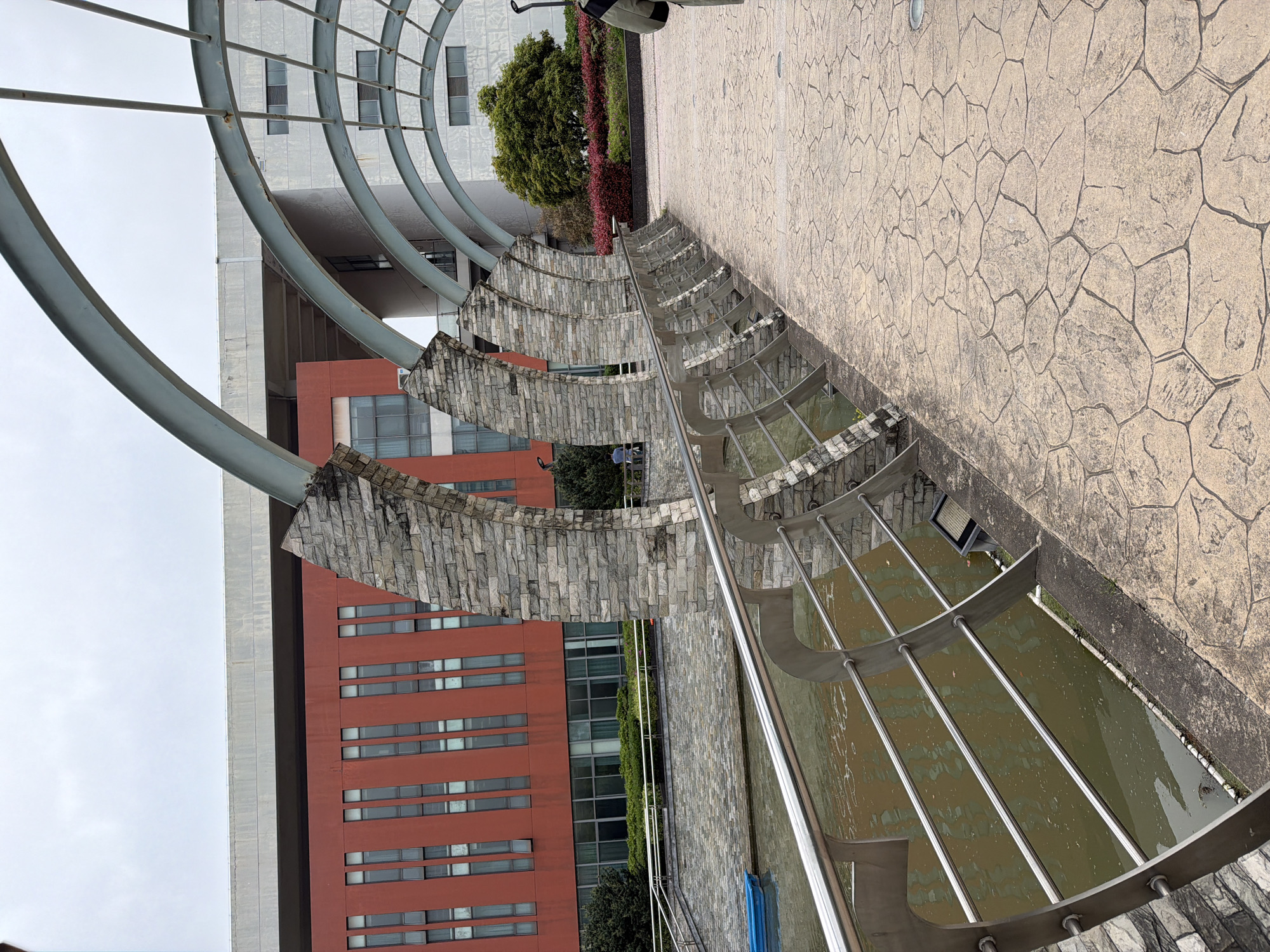 The photograph depicts an outdoor scene with distinct architectural and landscaping elements. Dominating the view are curved structures made of stone and metal, forming an arcade-like feature over a body of water. The water appears murky. Adjacent to this structure is a walkway paved with a cracked-stone pattern. Buildings are visible in the background; one is tall, with evenly spaced windows and a facade of reddish-brown color. Another building is gray, featuring curved architectural elements at the top. Foliage, including trees and flowering plants, is visible between the buildings and the water feature. Based on the location information (Jiangning District, China), the structures and architecture appear modern and possibly part of a park or public space.