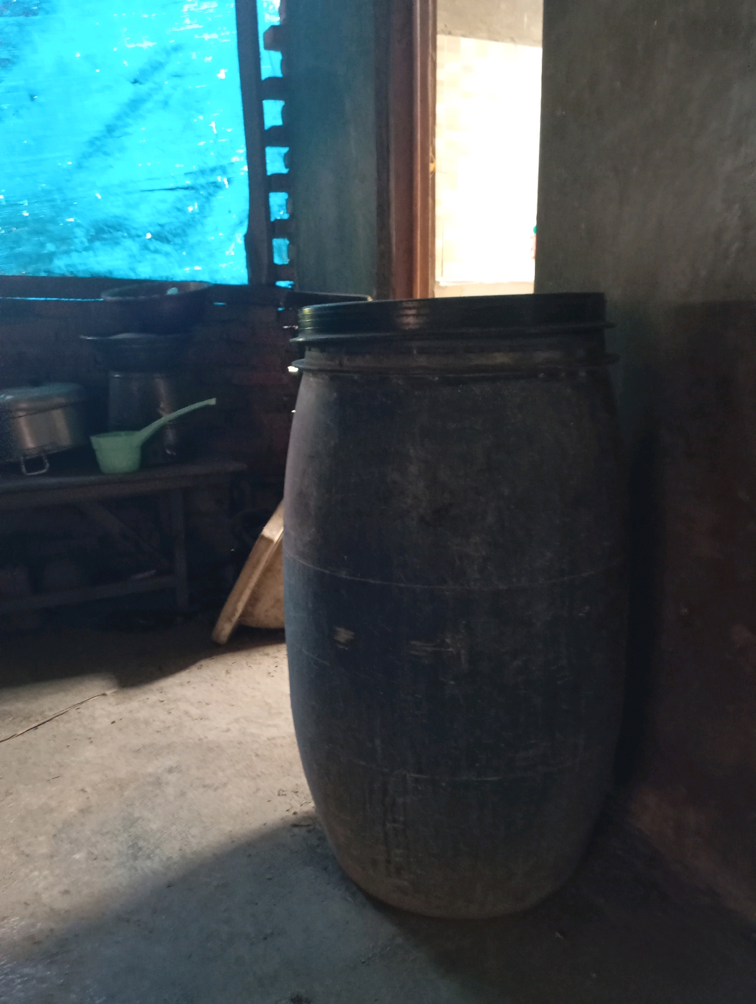 A large, dark-colored plastic barrel stands in the foreground of a dimly lit room, its surface showing signs of wear and dust. To the left of the barrel, a rustic wooden shelf holds various kitchen items, including a metallic pot with a lid and a pale green ladle. Behind the shelf, a blue tarp is visible, partially covering a window with visible wooden slats. The floor is concrete, with shadows cast by the barrel and other unseen objects. A wooden doorframe with a bright opening is visible to the right, suggesting an adjacent area. The overall impression is of a utilitarian space, possibly a kitchen or storage area in a rural or simple dwelling in Kecamatan Haurgeulis, Indonesia.