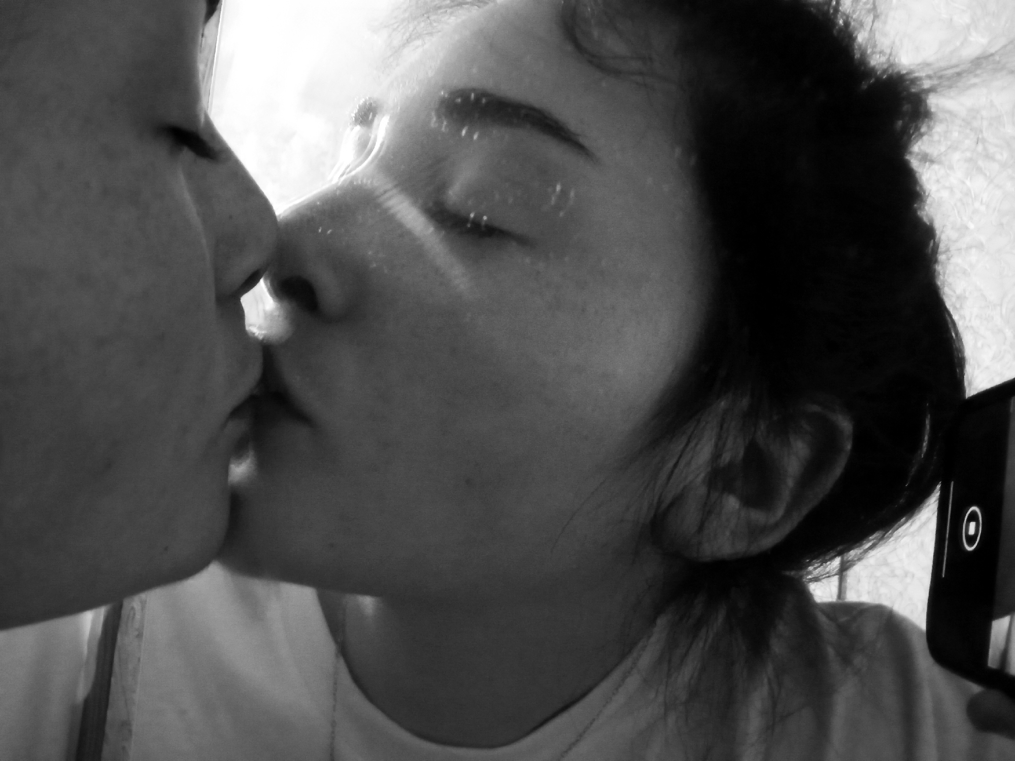 A close-up, black and white photograph depicts two individuals kissing. The person on the left has their face turned to the right, while the other person, whose face is angled to the left, has their eyes closed. A smartphone is visible in the lower right corner of the frame. The location in Poltava, Ukraine is not discernible from the image.