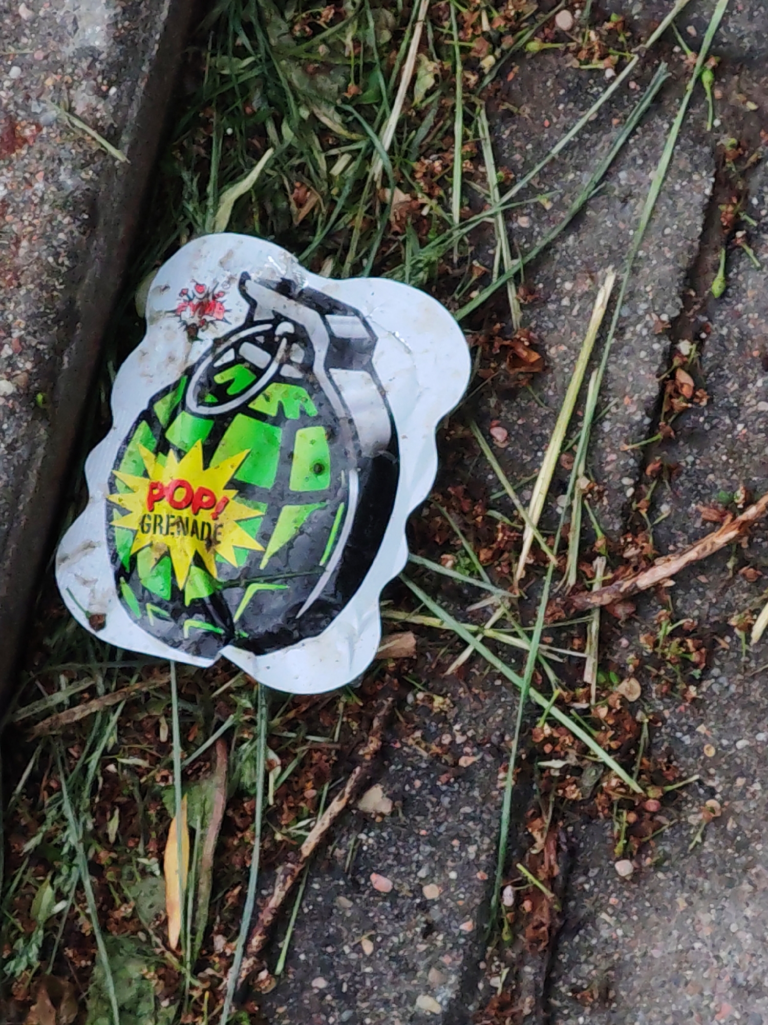 A discarded wrapper for a "Pop! Grenade" ice cream treat lies on a paved surface with grass growing through the cracks, next to a metal curb. The wrapper is white with a cartoon graphic of a green and black grenade. The words "Pop! Grenade" are printed on a yellow starburst in the center of the grenade image. There are reddish stains in the upper-left part of the wrapper. The pavement is grey and speckled with small stones and organic debris. The grass is green and brown.