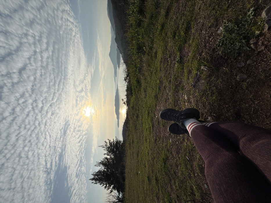 A first-person perspective photograph captures the lower legs of an individual resting at an elevated viewpoint in Vojnići, Montenegro. The subject's legs are extended and crossed at the ankles, clad in maroon ribbed leggings, white crew socks with two maroon horizontal stripes, and black hiking boots. The immediate foreground consists of sparse green grass, brown soil, and scattered small rocks. The midground features a grassy hillside, with a cluster of dark green pine trees visible on the left. The background presents a wide coastal panorama: a sun low on the horizon, positioned left of center, casts a bright, shimmering reflection across a body of water, likely a bay or the sea. Distant islands or landmasses are visible within the water, and undulating hills or mountains recede into the hazy distance on the right. The sky is largely covered by a textured pattern of numerous small, uniform cirrocumulus clouds against a pale blue expanse. The lighting suggests either sunrise or sunset.