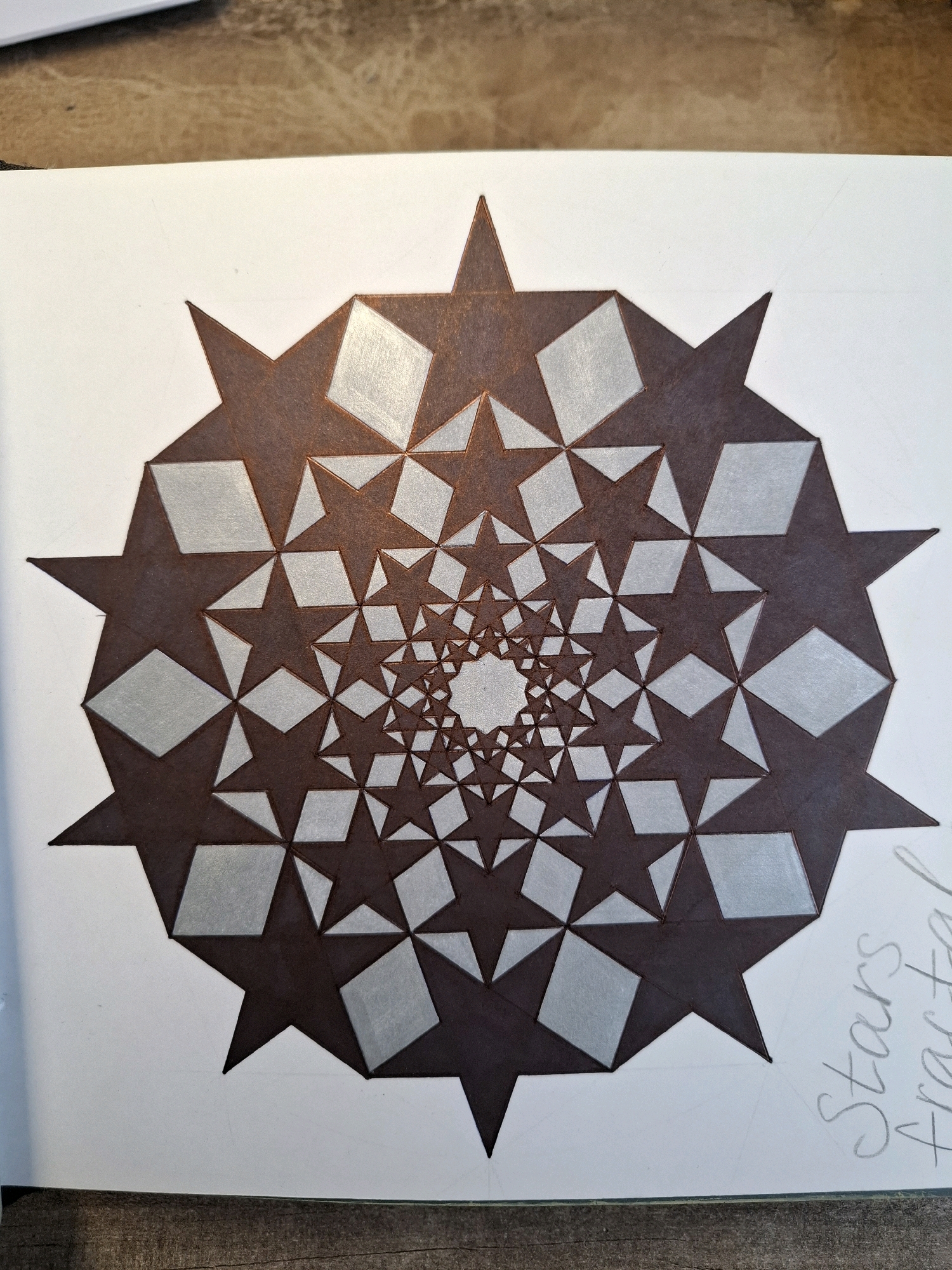 A close-up, top-down shot captures a drawing of a fractal star pattern on white paper. The pattern, rendered in dark brown and silver, spirals inward towards a central point. The outer layer features large, sharp-pointed stars alternating with diamond shapes. As the pattern progresses inward, the stars and diamonds become progressively smaller, with the stars appearing to be 5-pointed and the diamonds being rhomboid. The drawing is framed by the textured surface of a wooden table. To the right of the drawing, the words "Stars Fractal" are lightly written in pencil.