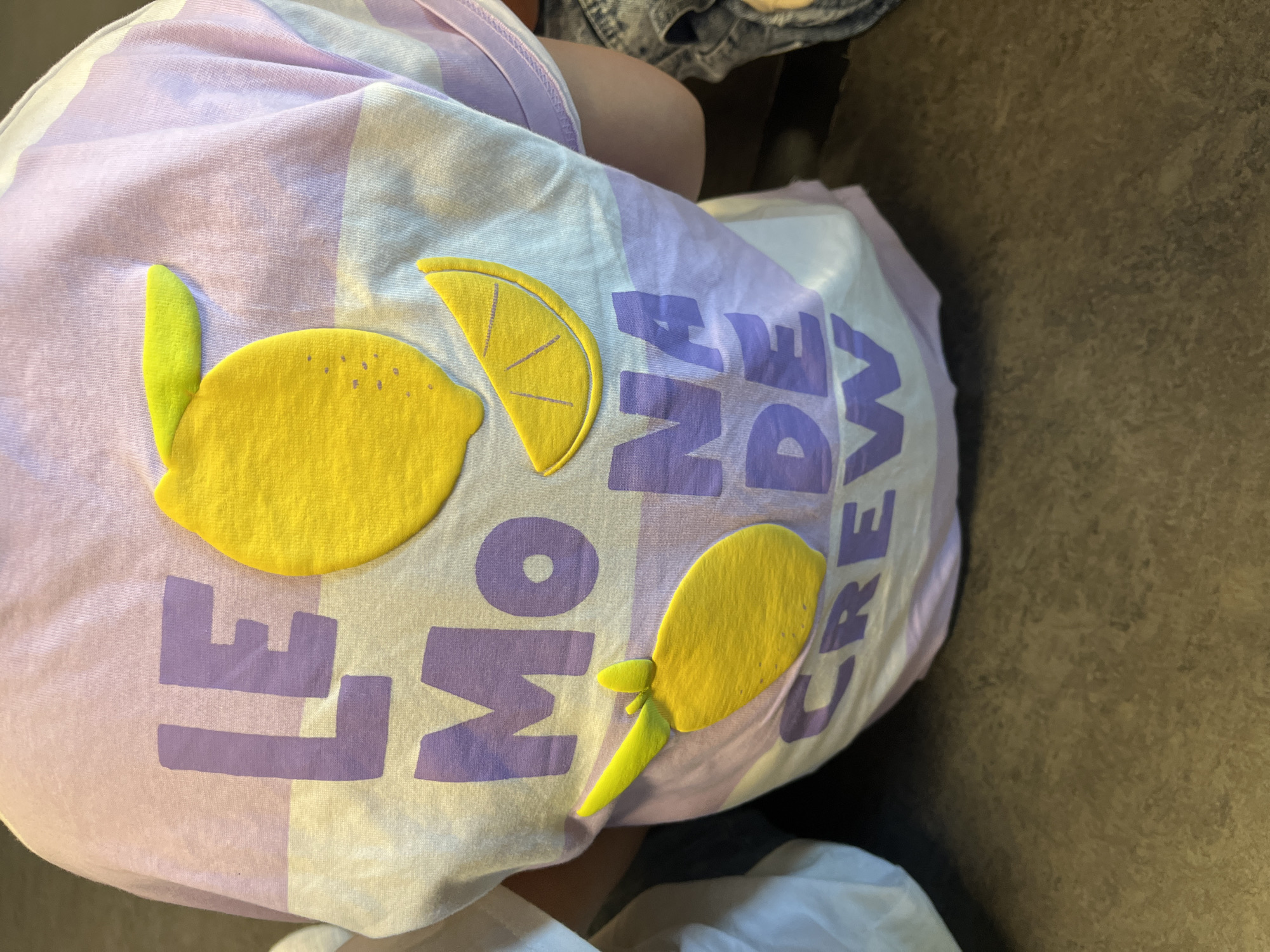 A close-up shot depicts a light purple and white checkered garment, possibly a shirt, adorned with stylized yellow lemon appliques and the text "LE MON DE CREW" in dark purple. The garment is situated on a neutral-colored surface, likely a floor or piece of furniture. A small portion of denim fabric is visible at the top of the frame. The location in La Fouillouse, France, is not discernible from the visual information.