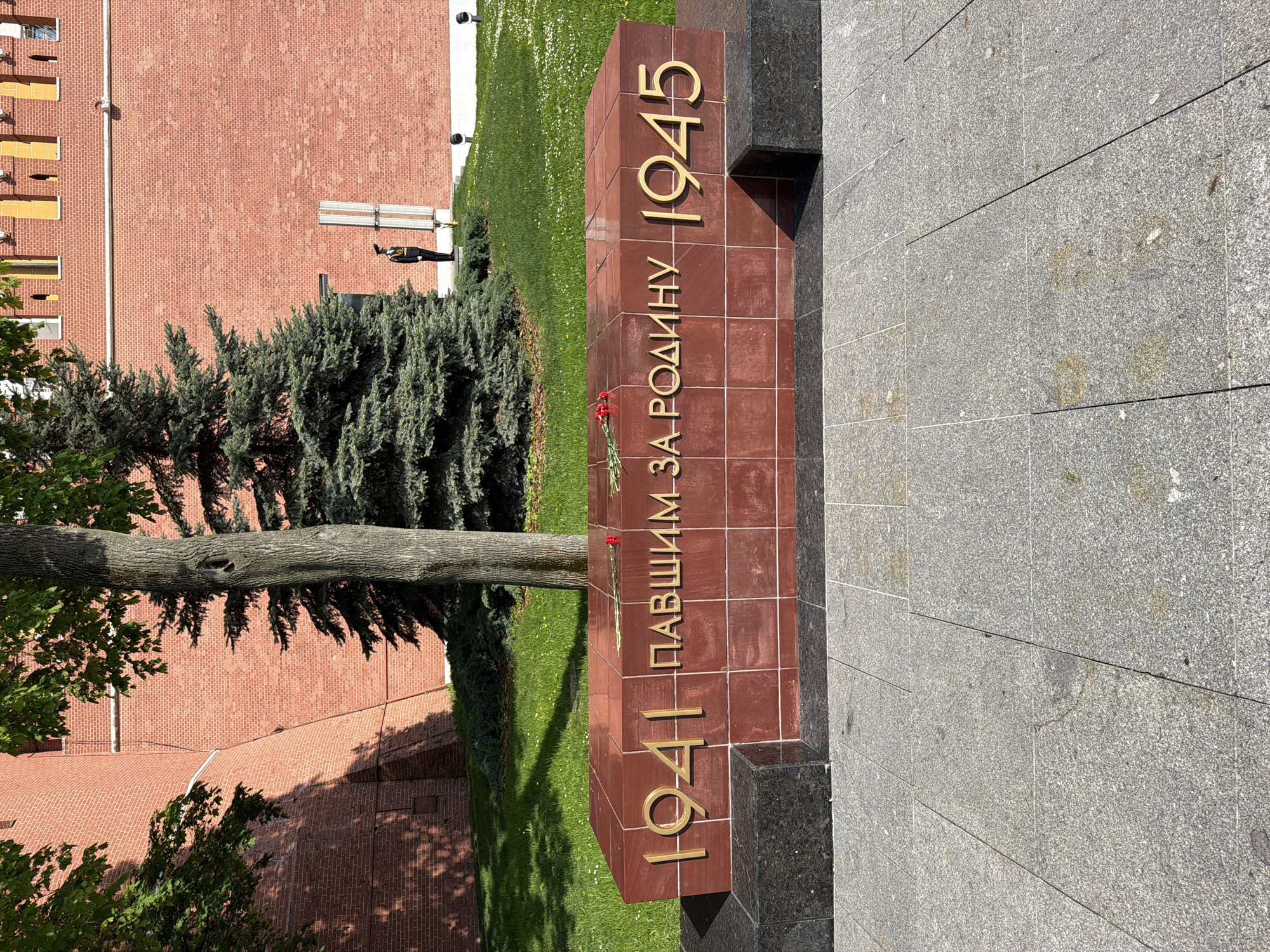 The image shows a monument in Moscow, Russia, likely located in or near Red Square. The monument is a rectangular prism made of reddish-brown stone, with the inscription "1941 ПАВШИМ ЗА РОДИНУ 1945" in gold lettering. The inscription is Russian and translates to "1941 To those who fell for the Motherland 1945". A few red flowers are placed near the center of the monument. The monument is flanked by black stone elements and sits on a gray stone platform. In the background, there is a green lawn, a spruce tree, and a brick building, partially obscured by foliage. Further in the background, a guard is standing by a platform. The scene is photographed during daylight with clear lighting conditions.