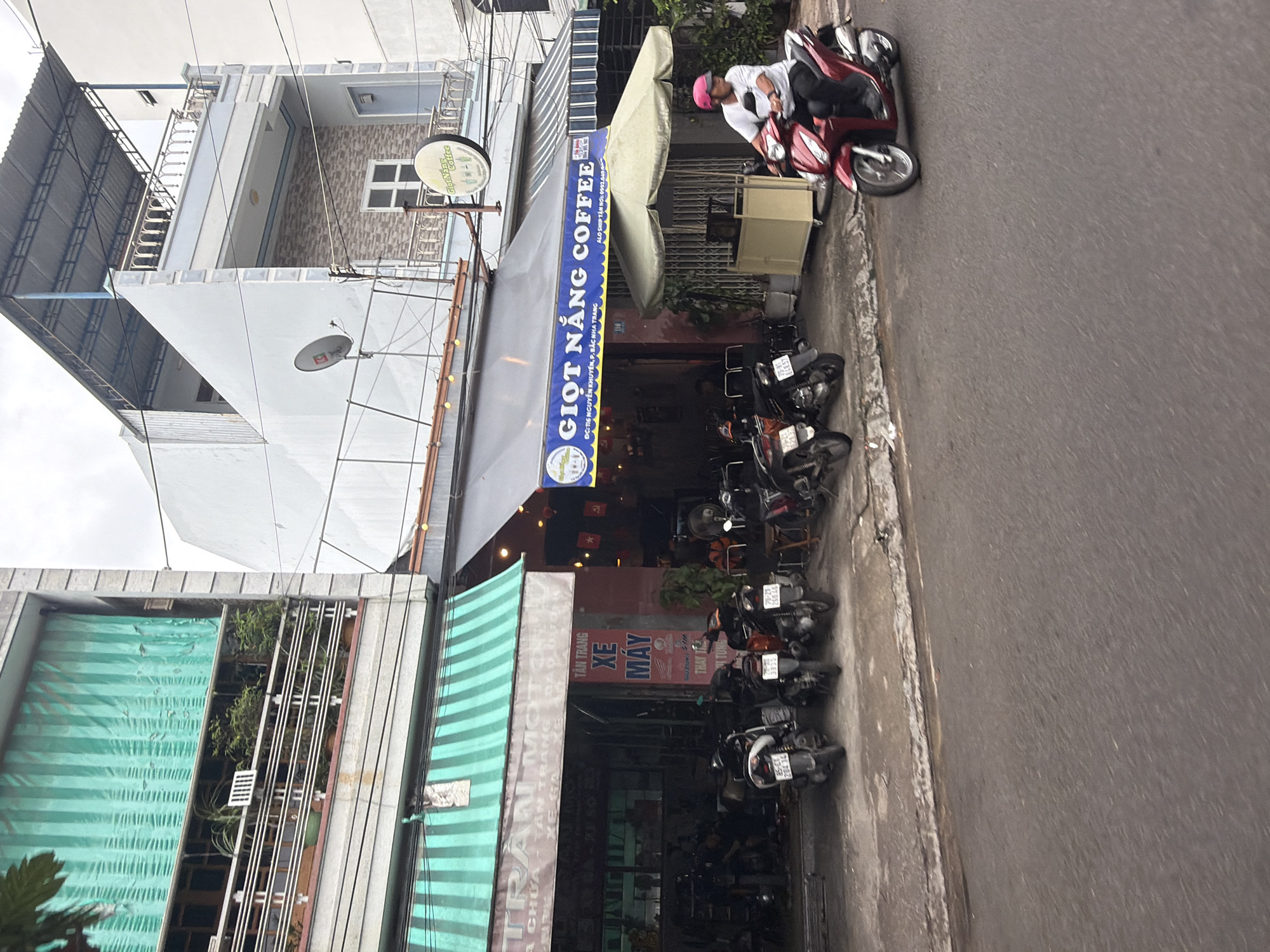 The image depicts a street scene in Nha Trang, Vietnam, with a focus on a row of buildings housing businesses. On the left, a building with green striped awnings features a motorcycle repair shop with several scooters parked outside. The sign above the shop reads "TÂN TRANG XE MÁY," indicating motorcycle refurbishment. To the right, a coffee shop with a blue awning displays a sign that says "GIỌT NẮNG COFFEE." The coffee shop appears to be a popular spot, with patrons visible inside. A person is riding a red motorcycle past the coffee shop. The street is paved, and the buildings are typical of urban Vietnamese architecture, with multiple stories and balconies. The sky is overcast, suggesting a cloudy day.