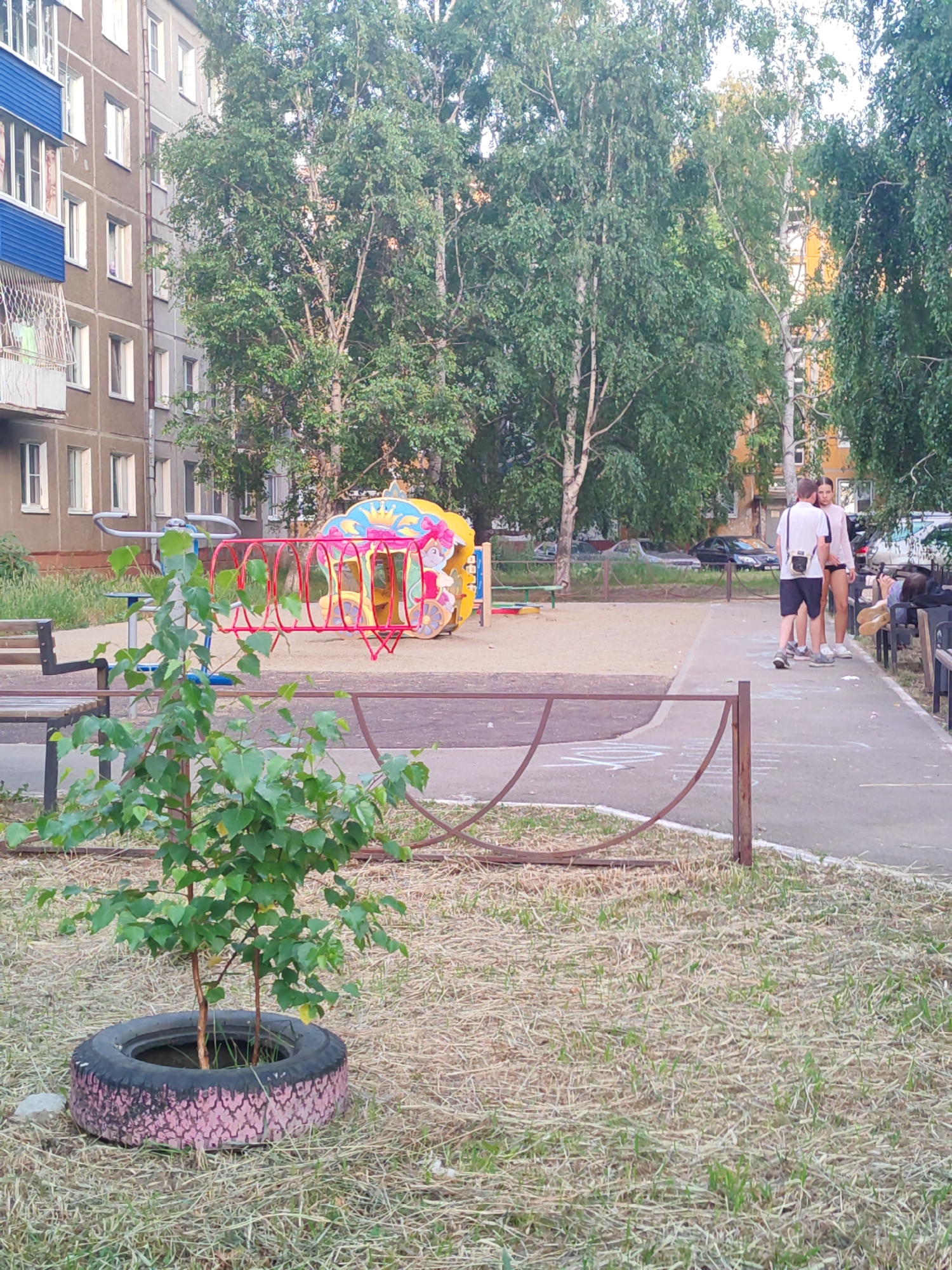 This is a medium-shot, eye-level photograph taken on a paved path next to a playground in Usolye-Sibirskoye, Russia. In the foreground, a small tree is planted in a tire that has been painted with a pink, mottled pattern. Behind the tree, a metal fence runs horizontally across the image, separating the playground from the grassy area. The playground itself features a colorful, circular climbing structure with red bars and a yellow and pink canopy depicting cartoon characters. To the right of the playground, a concrete path is visible, with two young adults standing on it, facing each other. The person on the left wears a white t-shirt and black shorts, carrying a black bag over their shoulder. The person on the right wears a light-colored top and shorts. Further to the right, another paved area with benches is visible, with several people seated. In the background, apartment buildings with balconies and trees line the area. The sky is overcast.